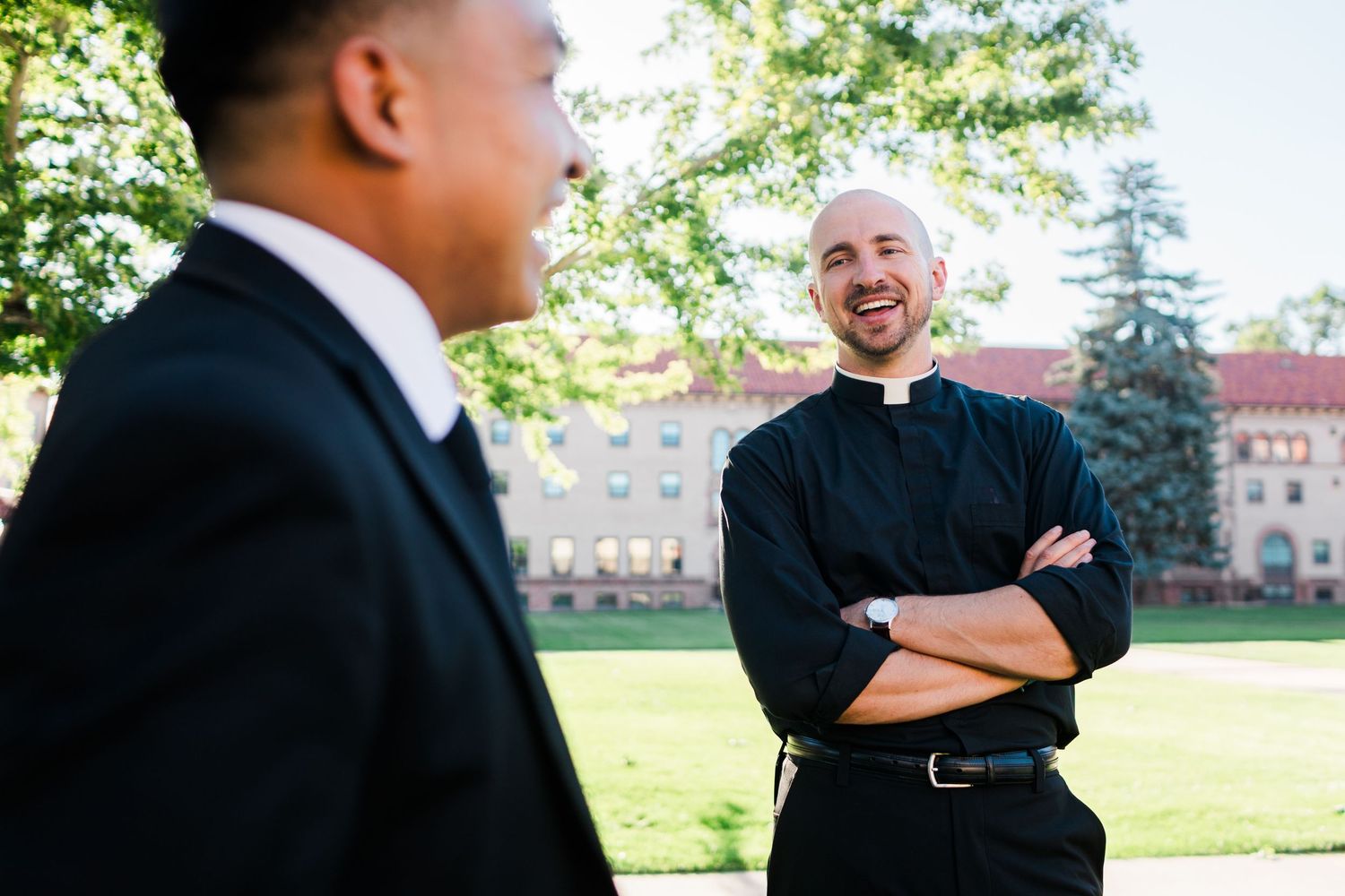 A smiling bald priest in black clergy attire stands with arms crossed outdoors, speaking with a man in a suit.