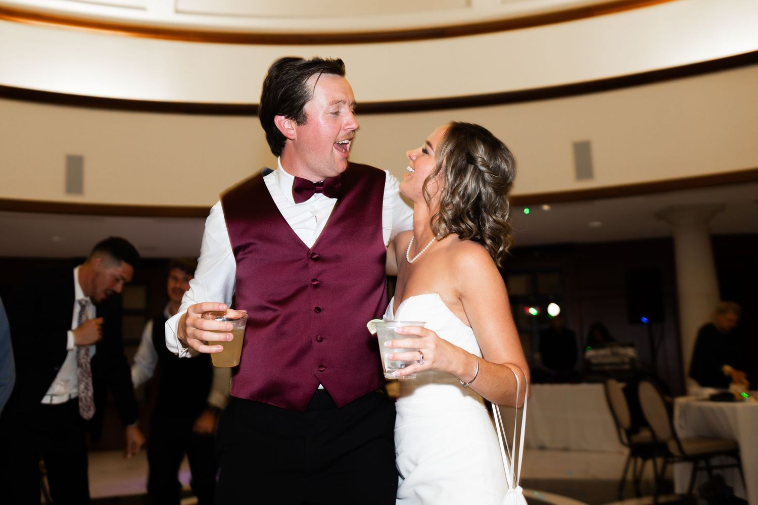 Bride in white strapless gown laughing with groom in burgundy vest and bow tie during wedding reception dance.
