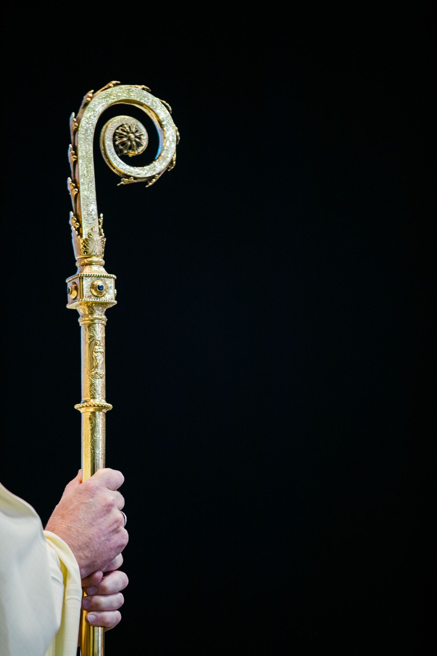 Golden bishop's crosier staff held by clergy in white vestments against dark background.