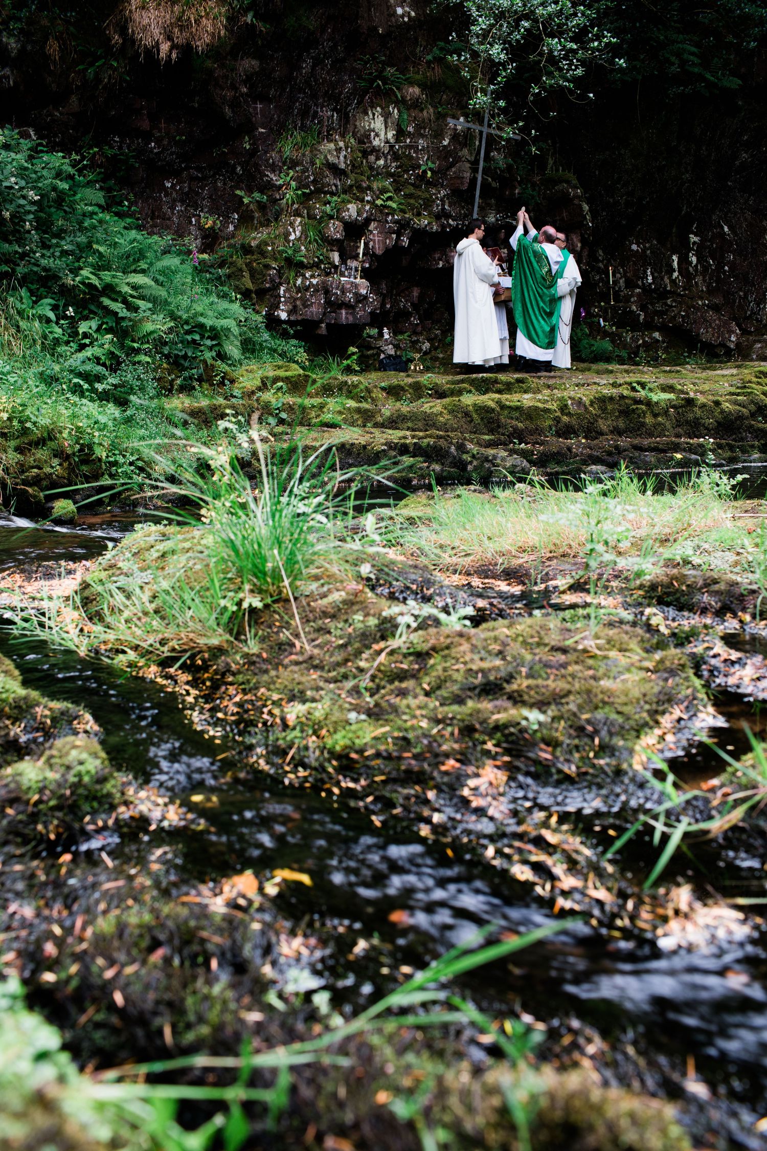 Two priests in white and green vestments performing an outdoor ceremony near a stream.