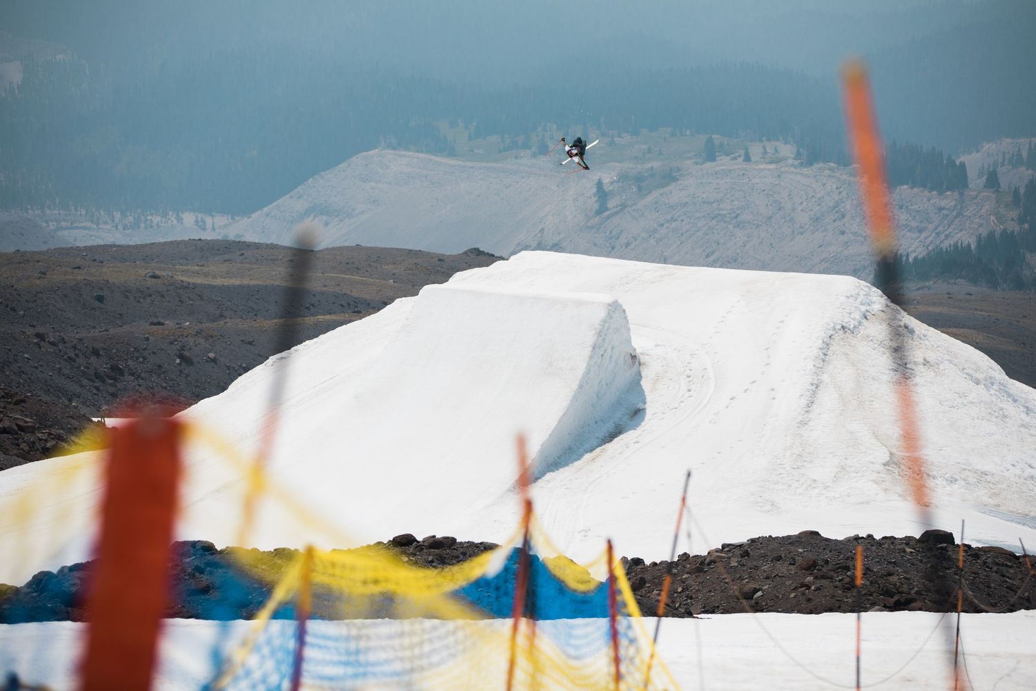 A distant skier jumps over a snowy slope with colorful flags and markers in the foreground.