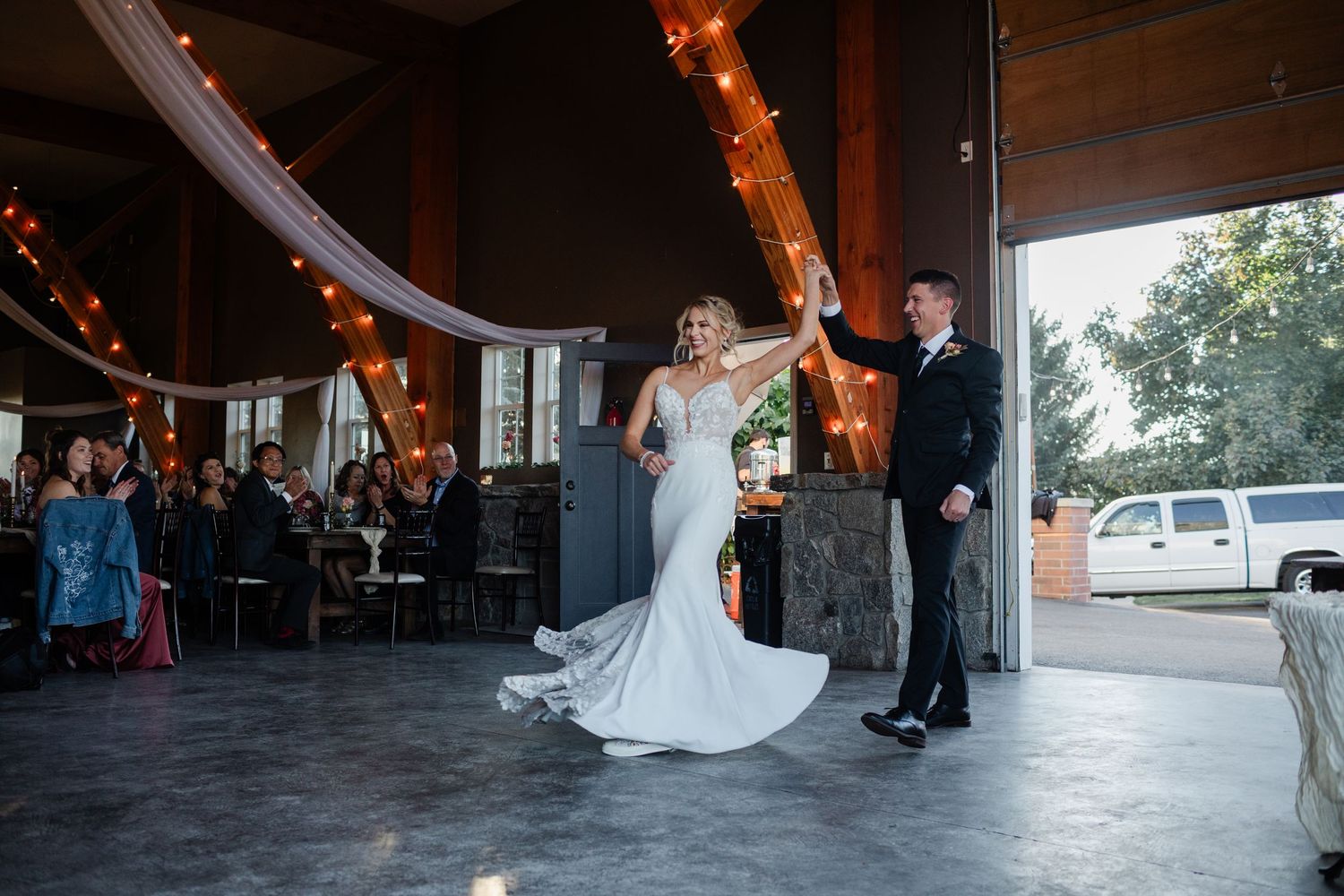 Bride and groom share their first dance under warm string lights in an elegant rustic wedding venue.