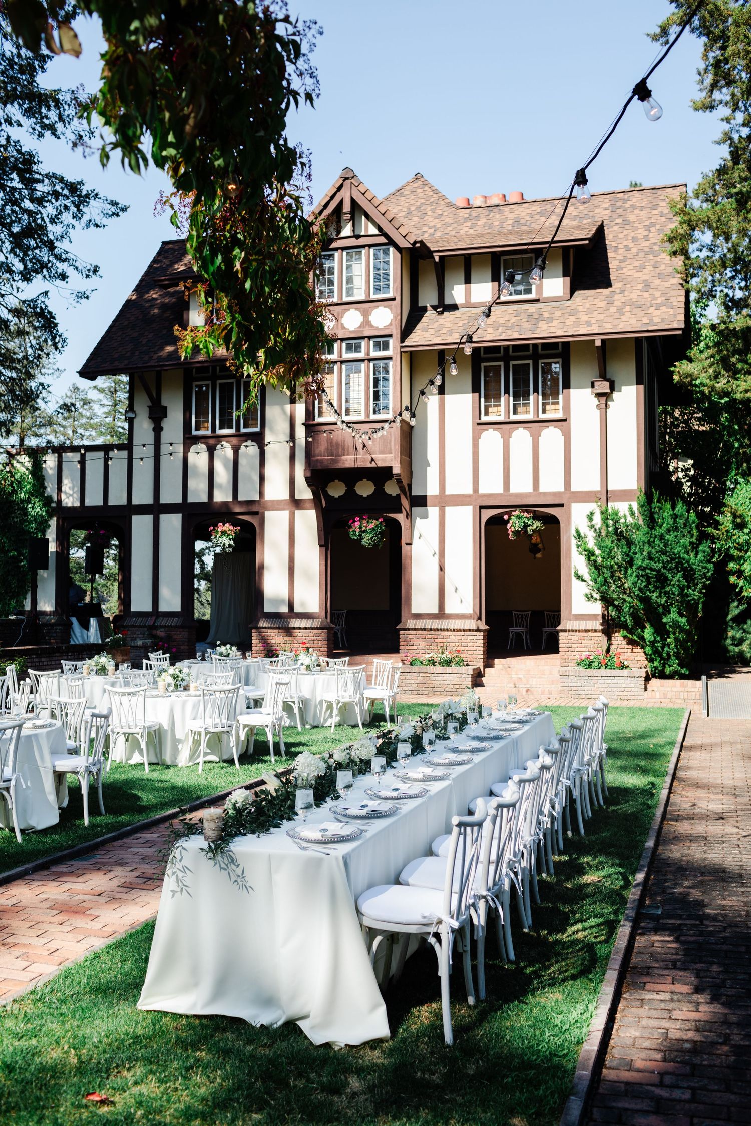 Elegant outdoor wedding reception with long white tables set on lawn before Tudor-style mansion.