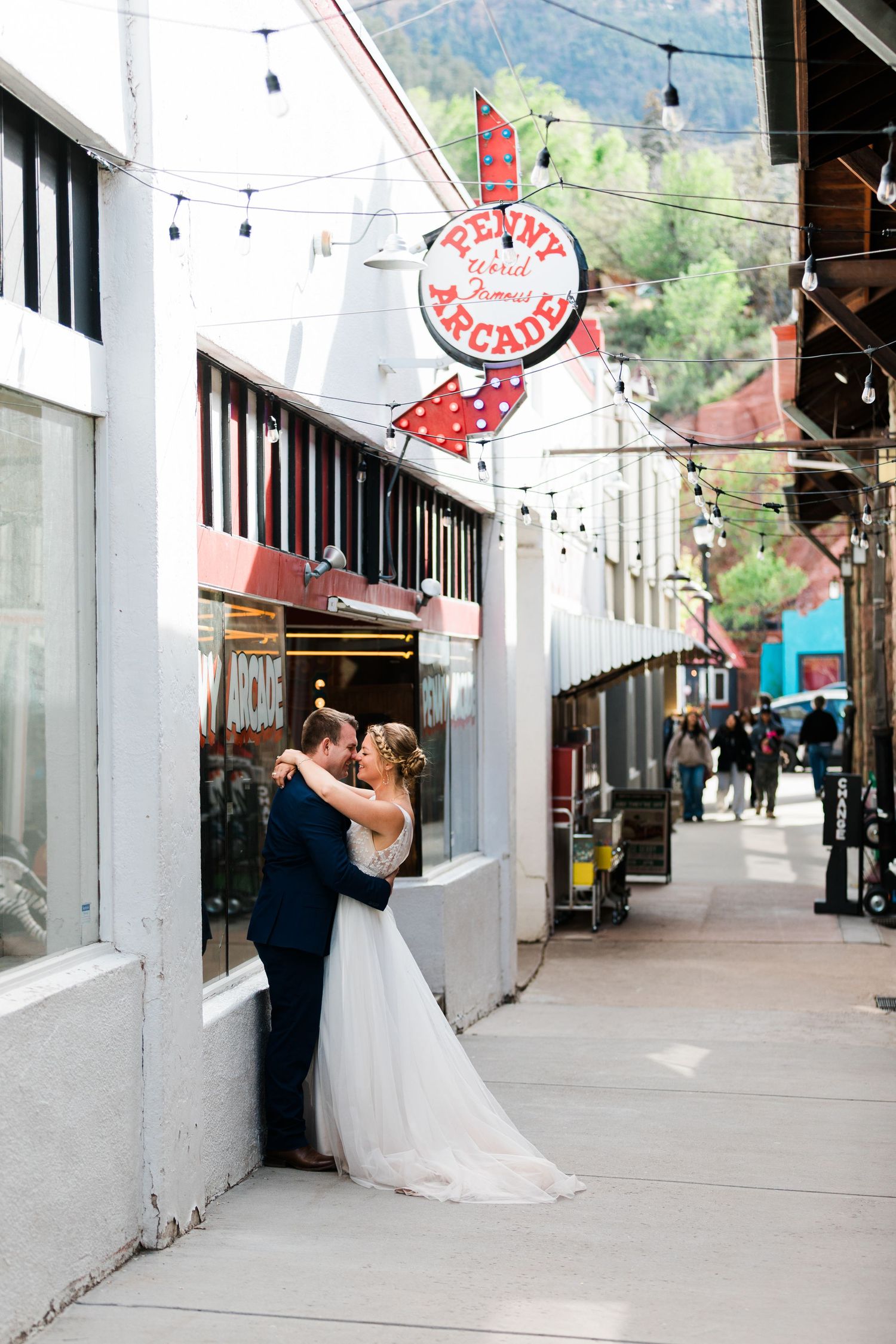 Bride and groom share a romantic embrace outside a Penny Arcade on a charming small-town street lined with string lights.