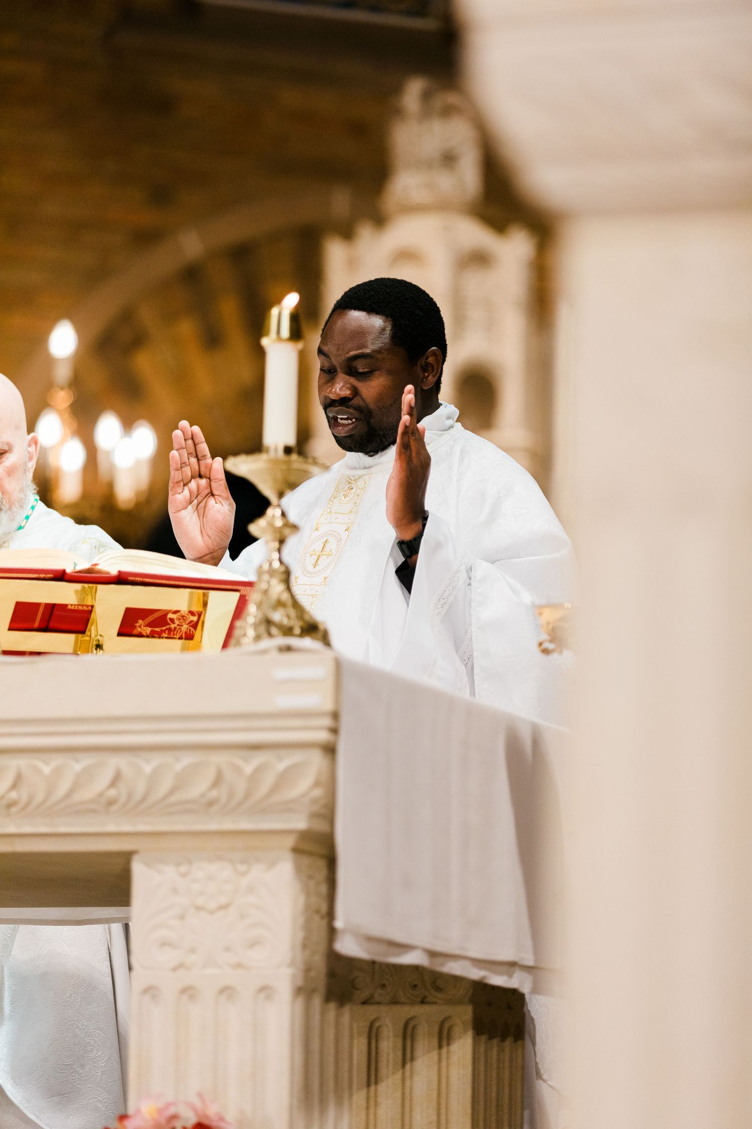 Black Catholic priest in white vestments raises hands in prayer at ornate church altar during Mass.