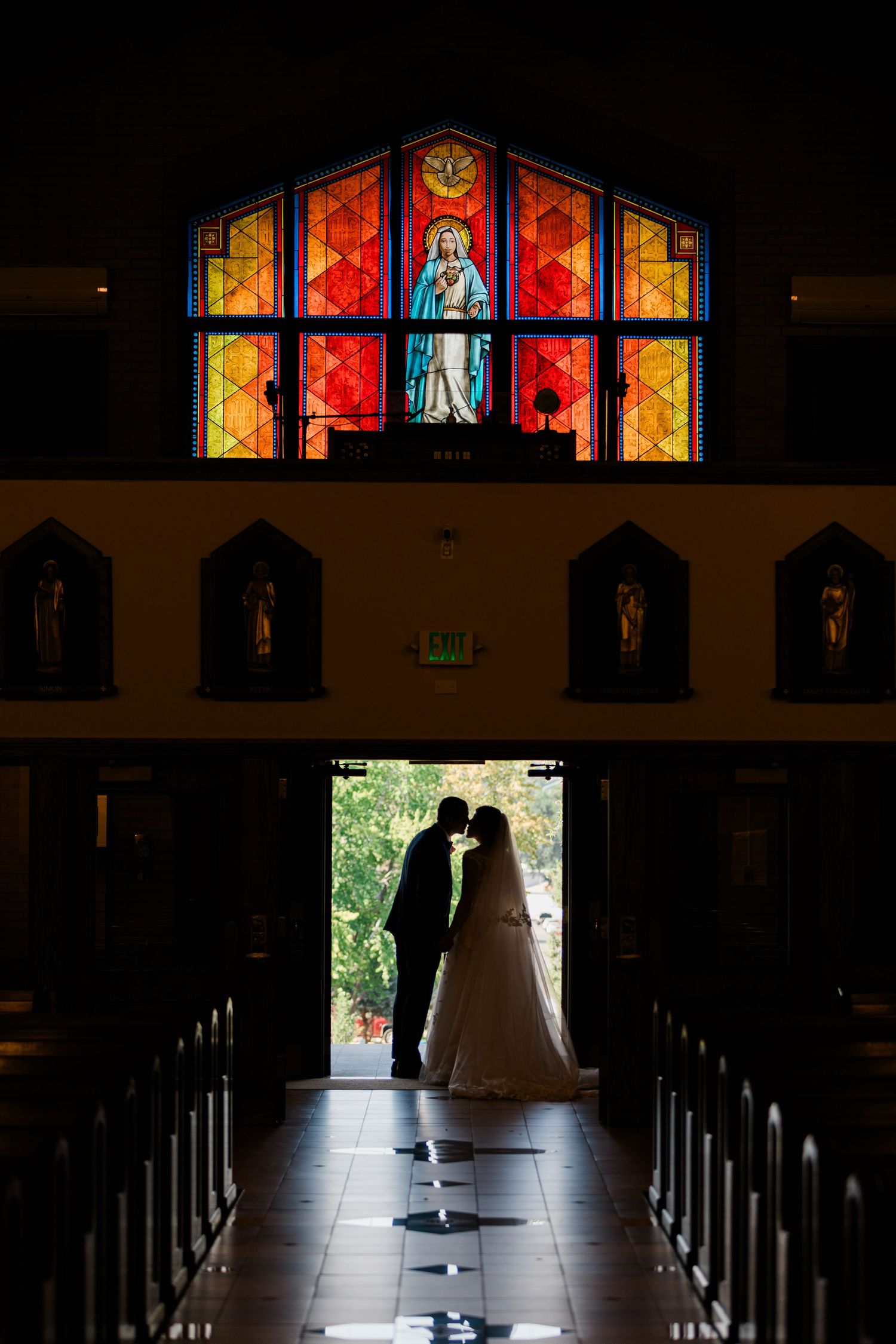 Bride and groom silhouetted kissing in church doorway with vibrant stained glass window of Virgin Mary glowing above.
