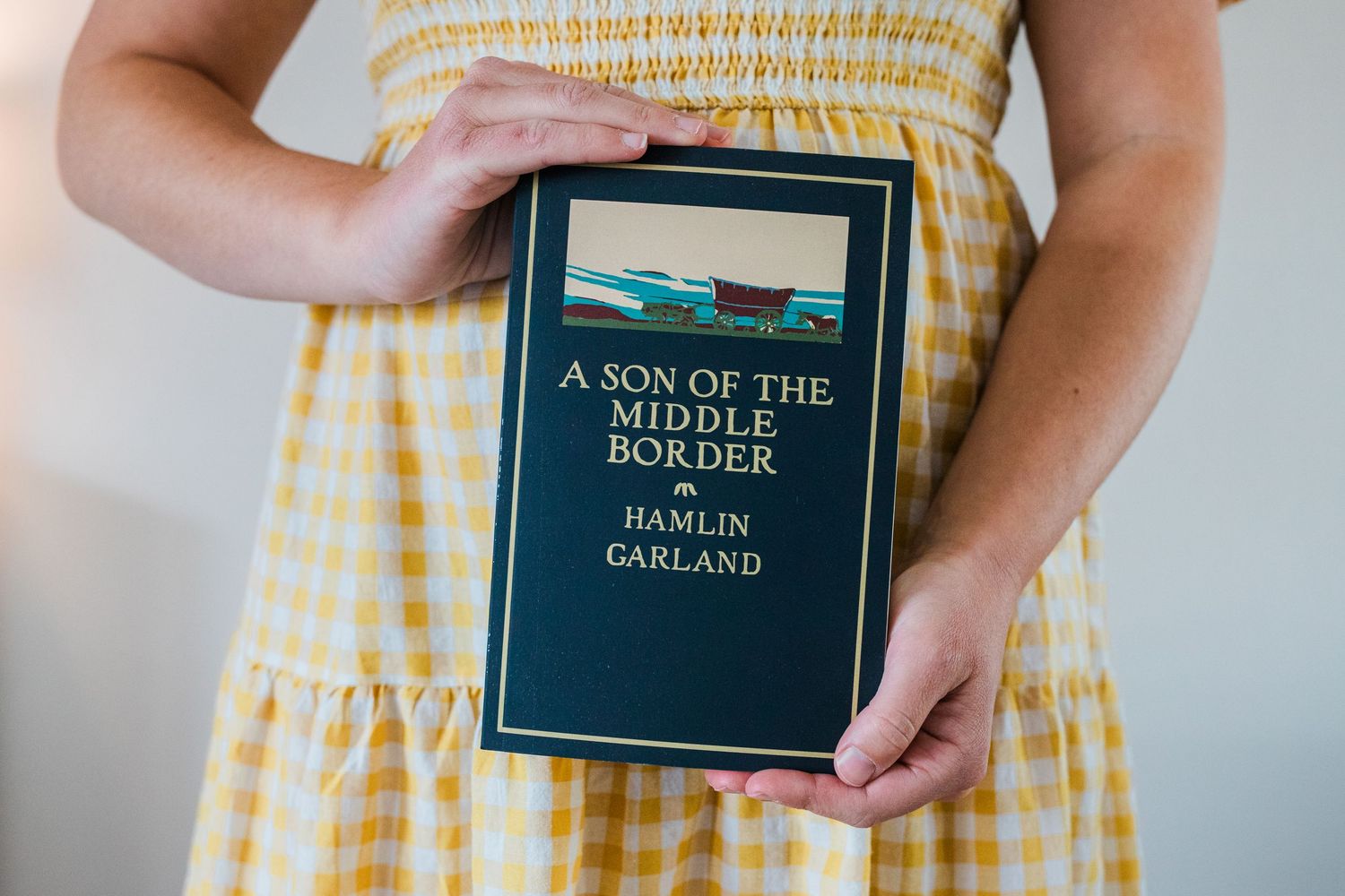 Woman in yellow gingham dress holds a small black book against a light colored wall.