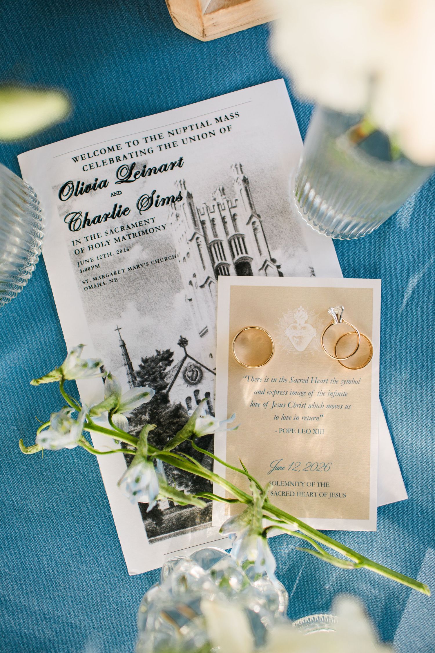 Wedding stationery flat lay featuring ceremony program, rings, and vows card on blue linen.