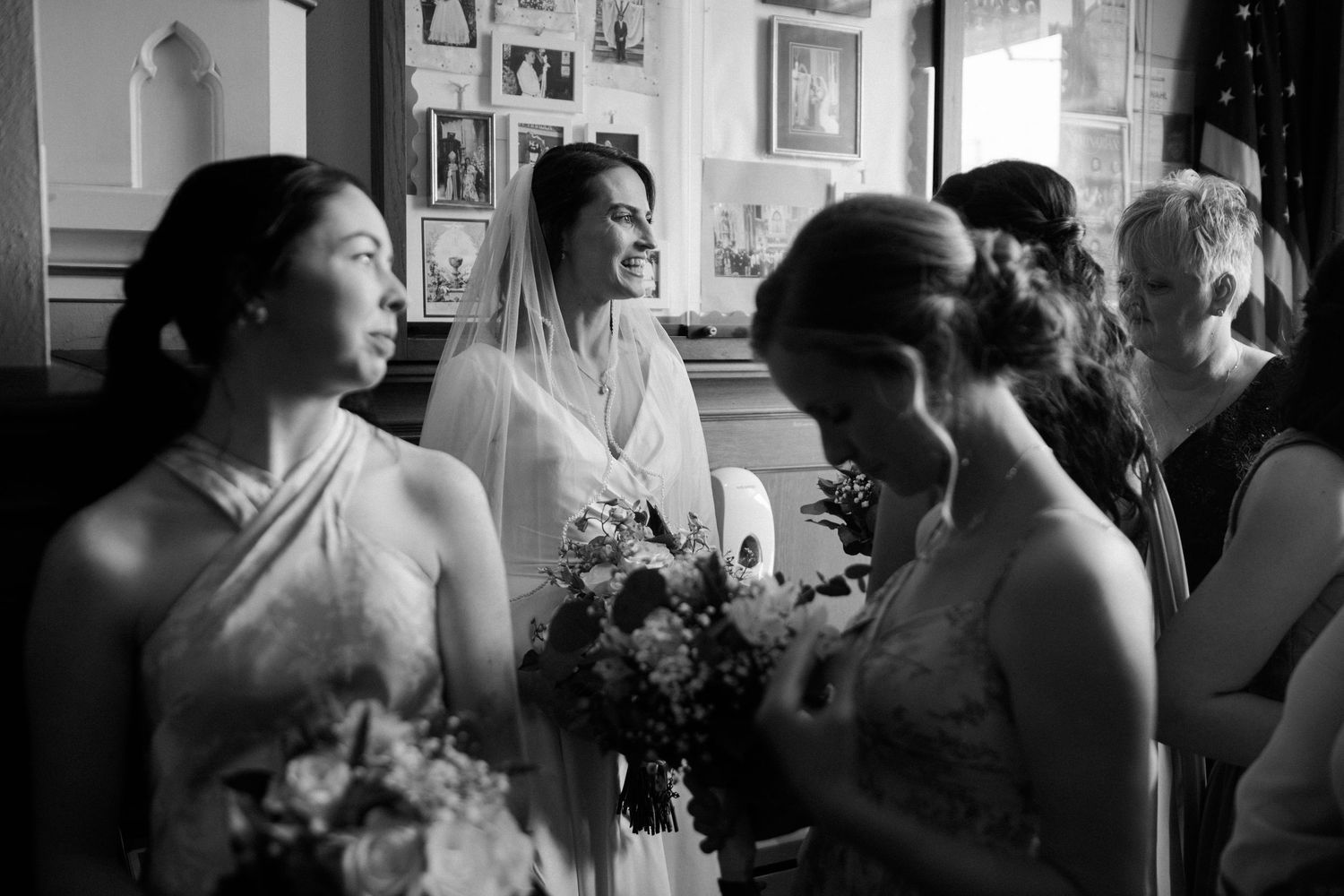 Black and white photo of a smiling bride holding a bouquet surrounded by bridesmaids at a wedding ceremony.