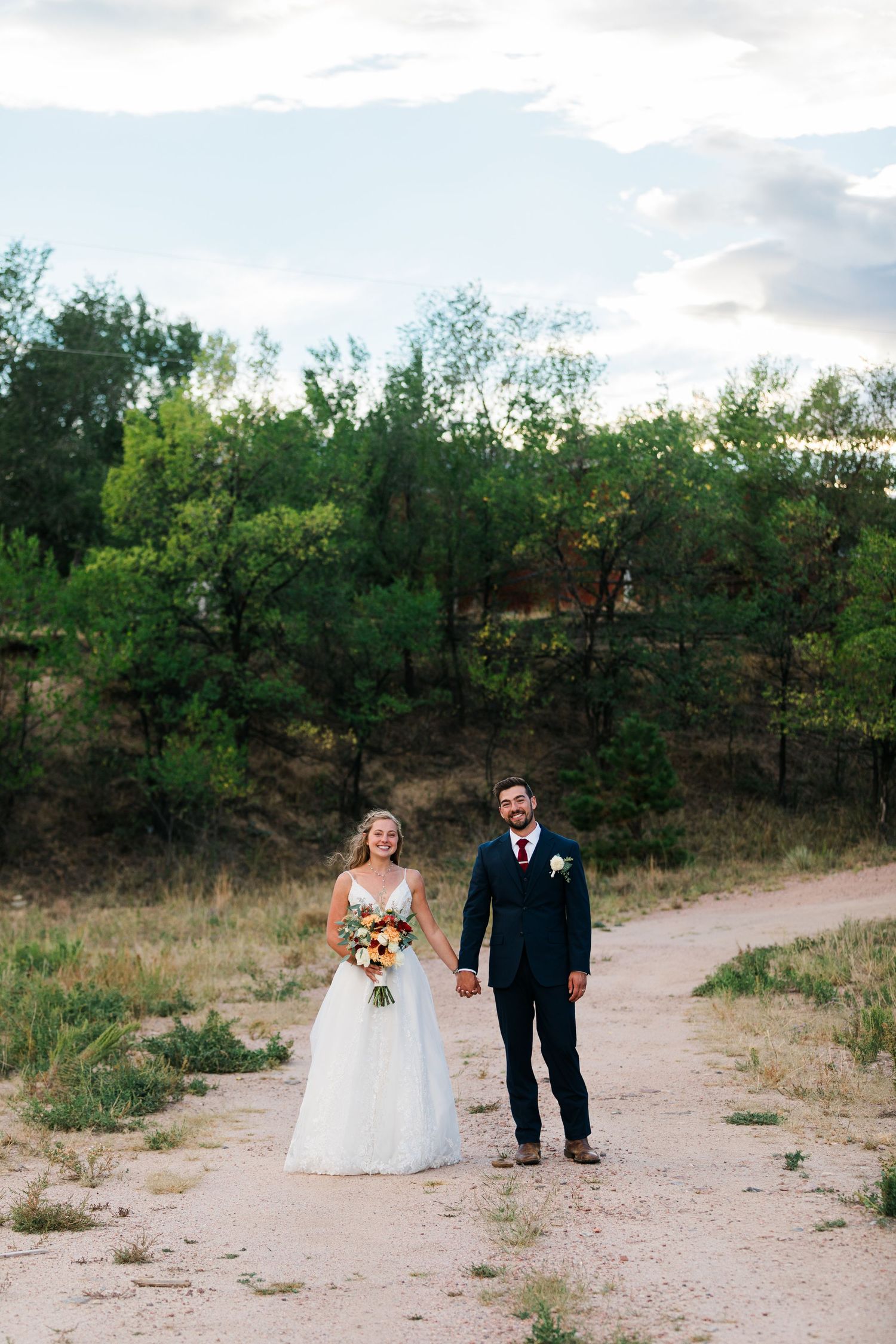 Bride and groom holding hands on a dirt path surrounded by lush green trees on their wedding day.