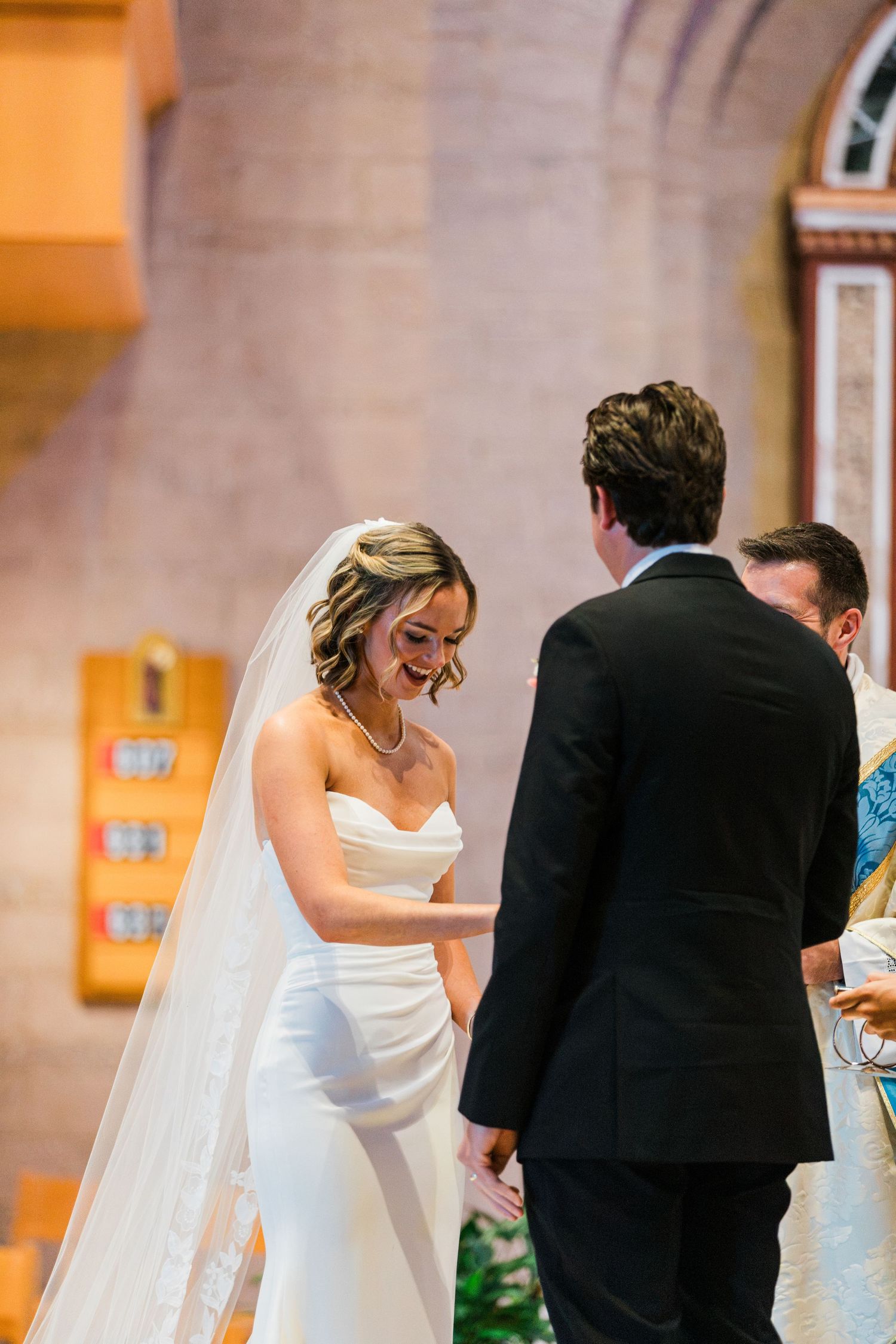 Bride in white gown and veil smiling during wedding ceremony at church altar with groom in black suit.