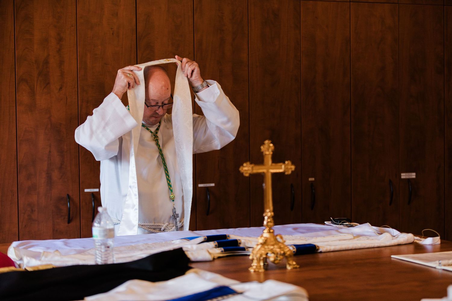 Priest in white vestments preparing for Catholic Mass at an altar with a gold cross.