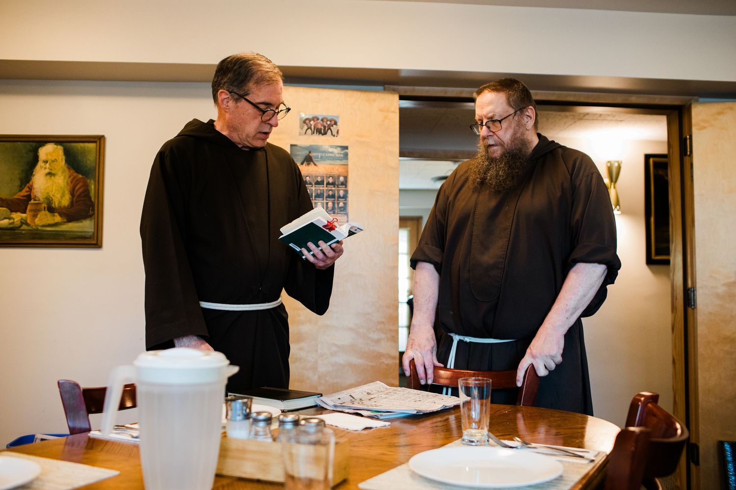 Two Catholic friars in brown habits reviewing a book at a dining table indoors.