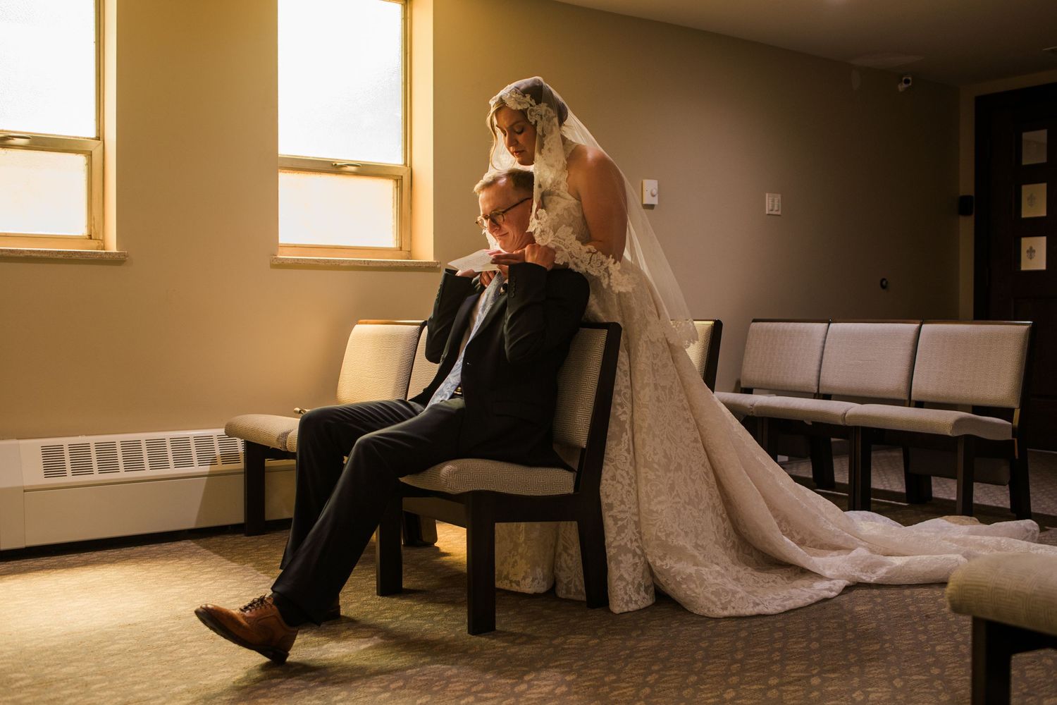 Bride in lace gown stands behind groom seated in chair during intimate wedding moment in softly lit room.