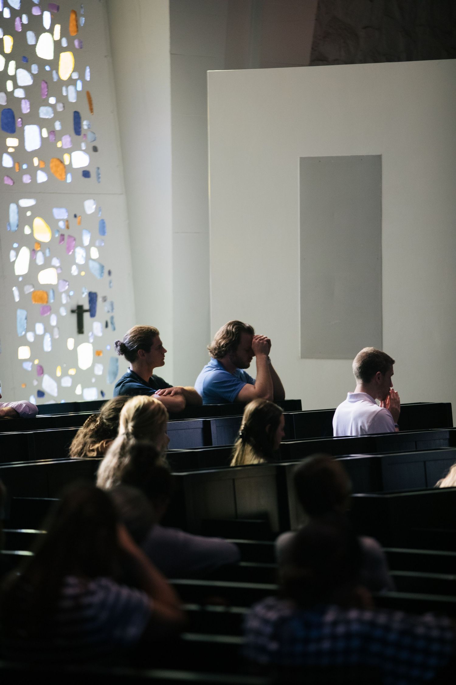 People sit in dark auditorium seating with colorful stained glass windows visible along the wall.