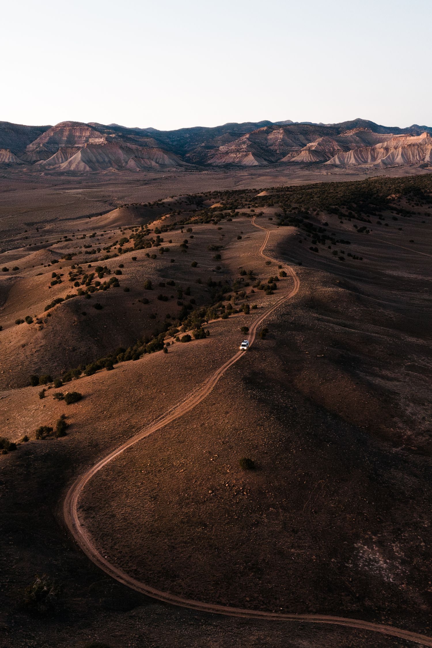 Winding dirt road curves through rugged desert landscape with mountains in the background during golden hour.