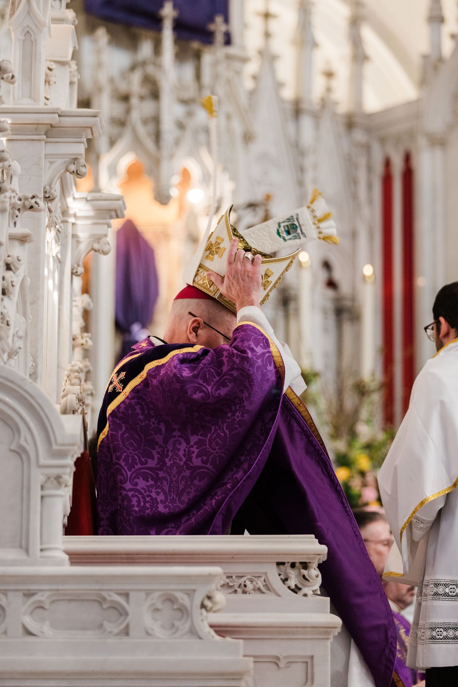 A figure in ornate purple robes and golden crown bows during a religious ceremony in an elaborate cathedral setting.