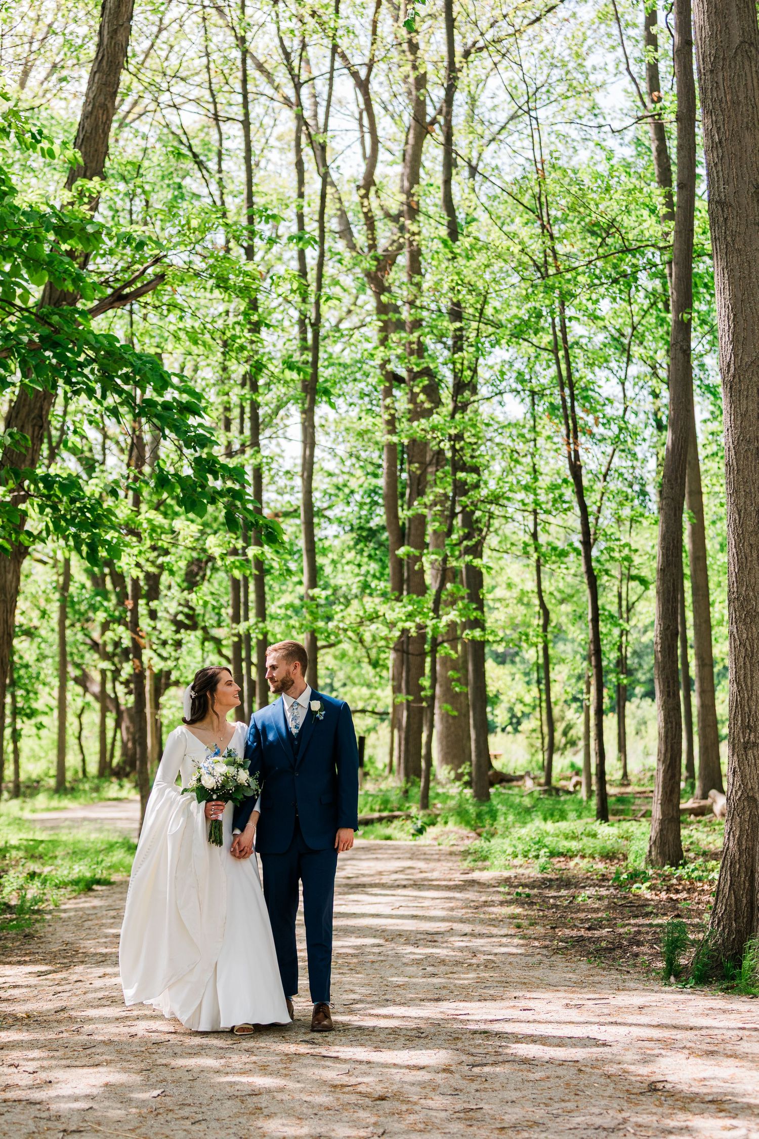 Bride and groom walk hand in hand along a tree-lined path in a lush green forest.
