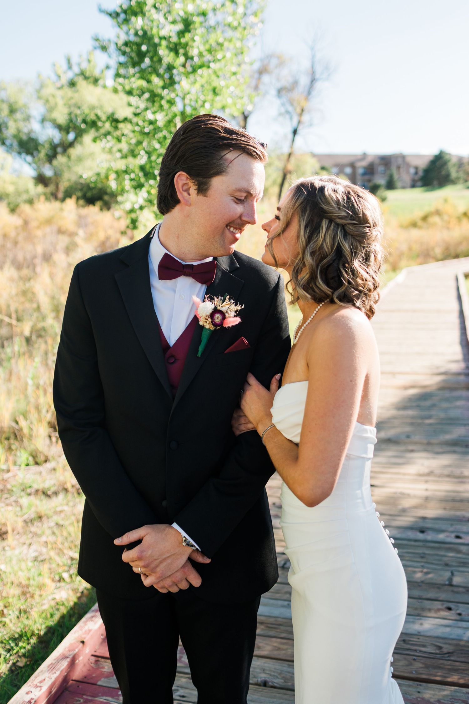 Bride and groom share romantic moment on wooden bridge, groom in black tuxedo with burgundy bow tie.