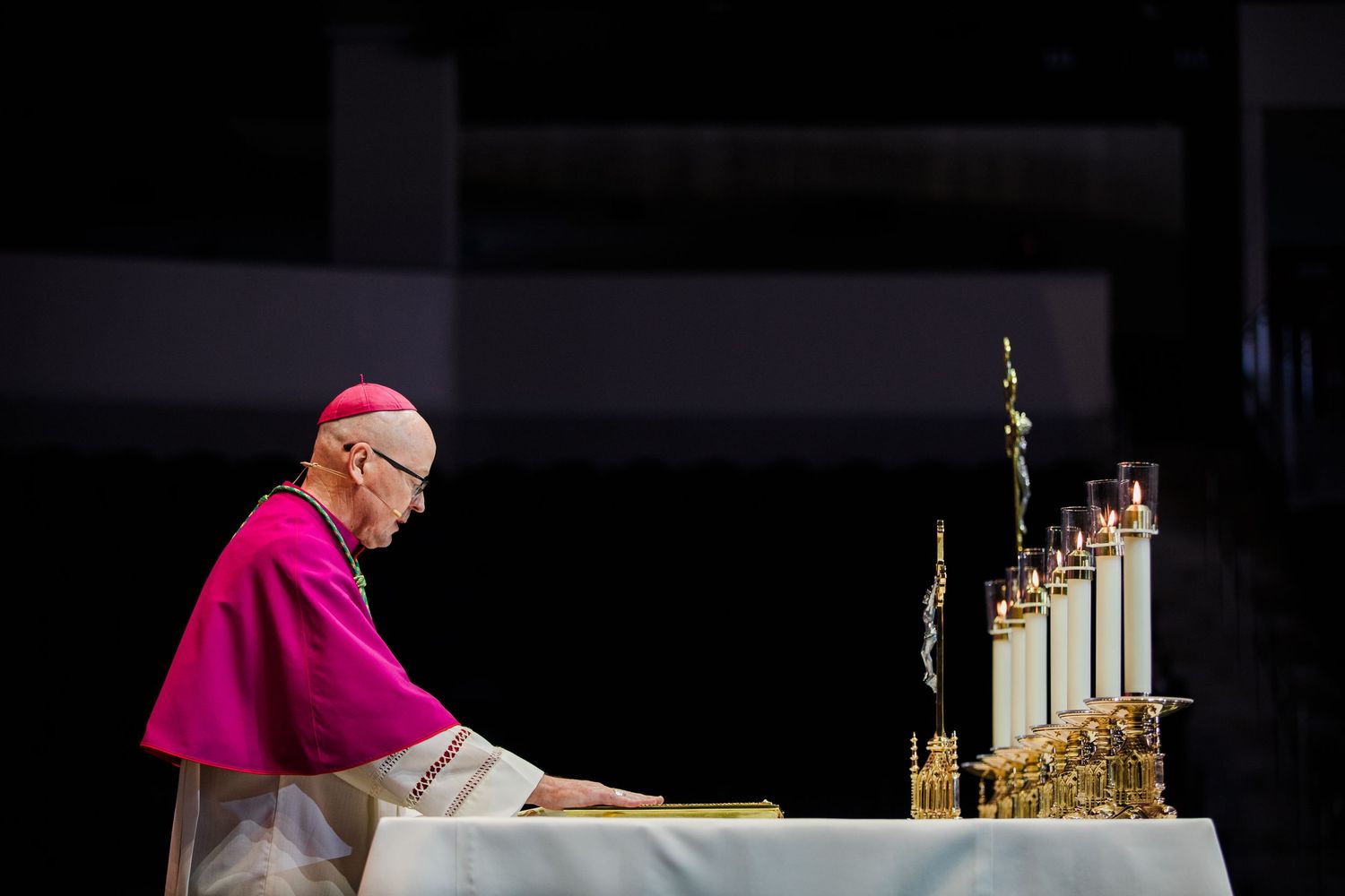 A bishop in magenta robes performs a ceremonial ritual at an altar with candles.