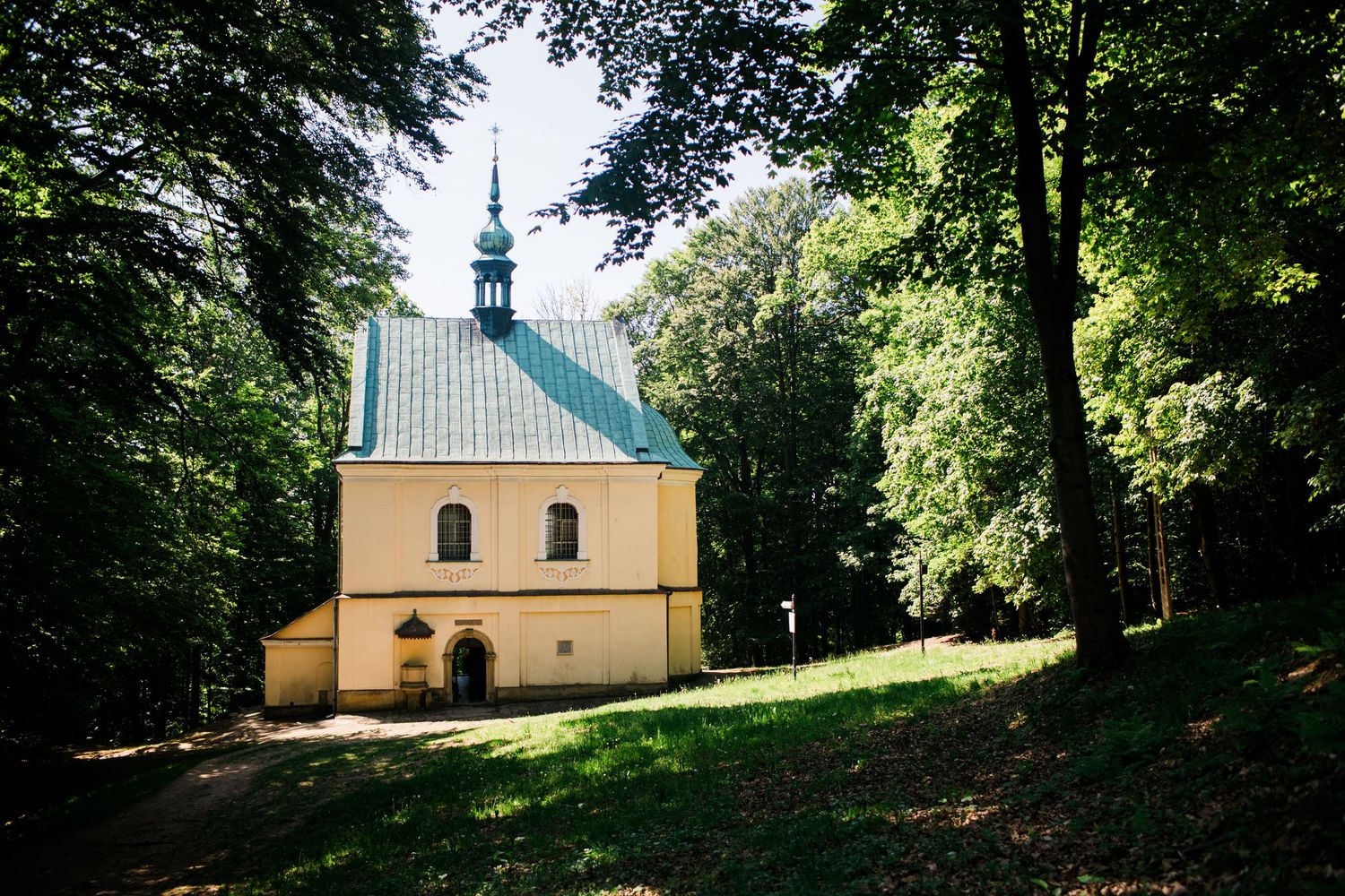 Yellow chapel with green roof nestled among tall trees in a sunlit forest clearing.