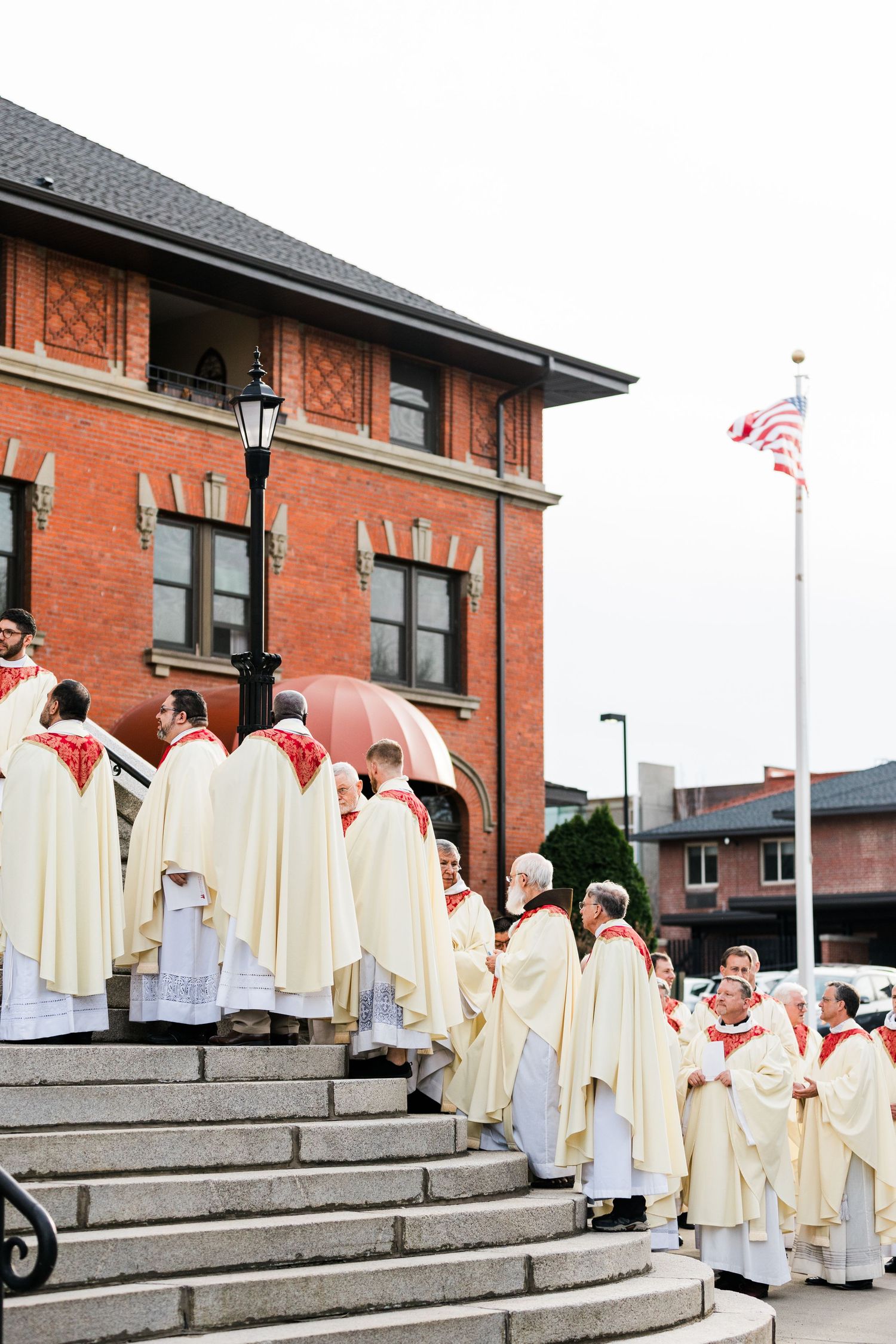 A line of clergy in cream-colored robes stand on the steps of a red brick building with an American flag.