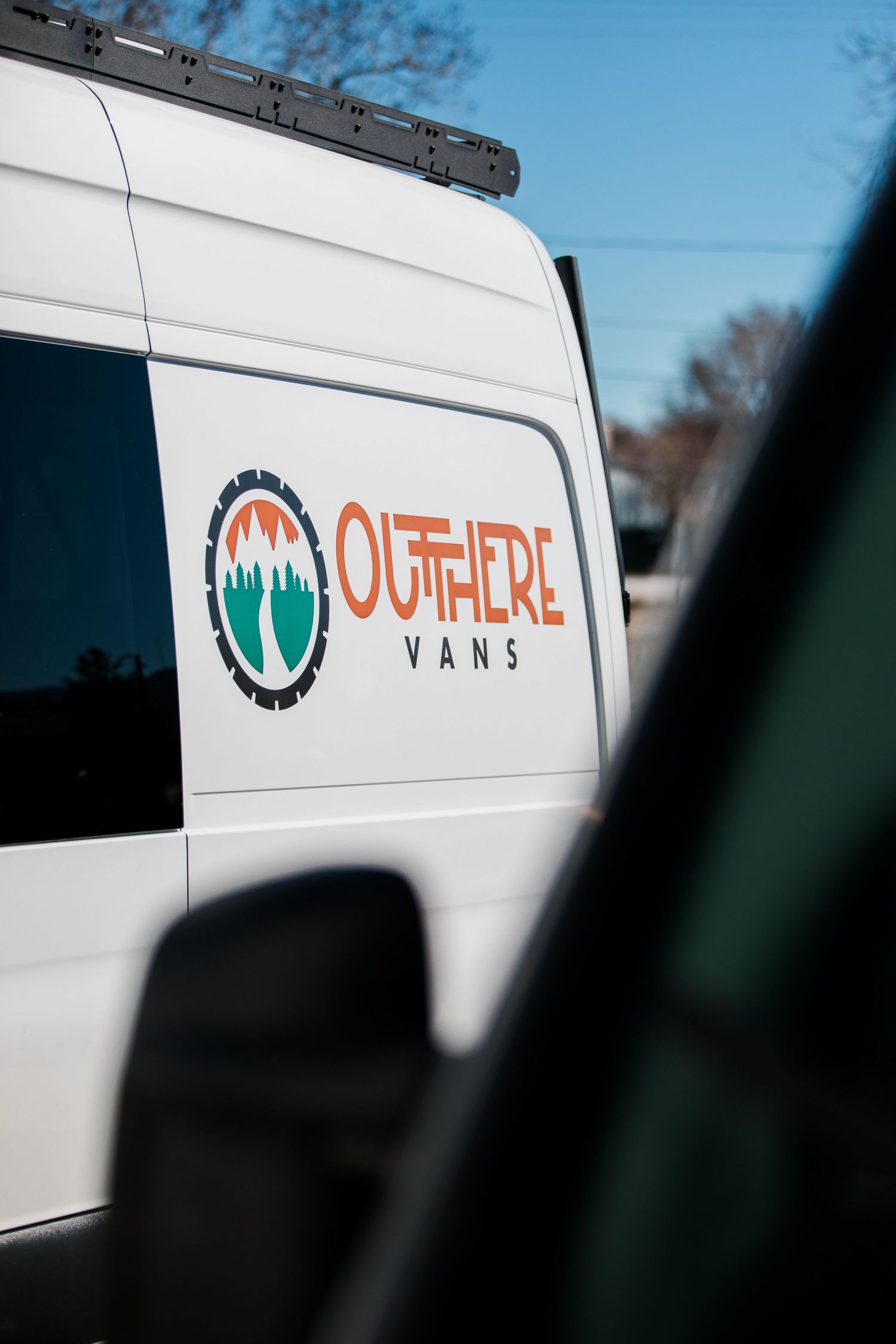 White van with Out There Vans logo and branding seen through a car window against blue sky.