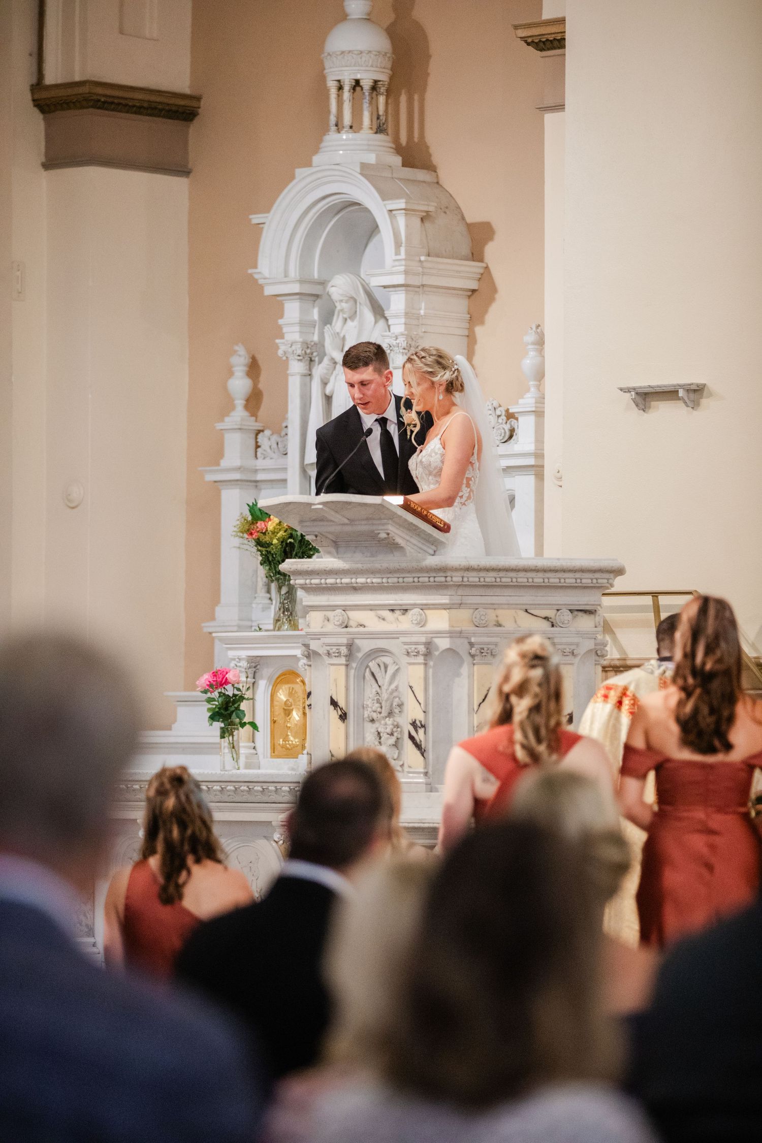 Bride and groom stand at ornate white church altar reading vows before seated wedding guests.
