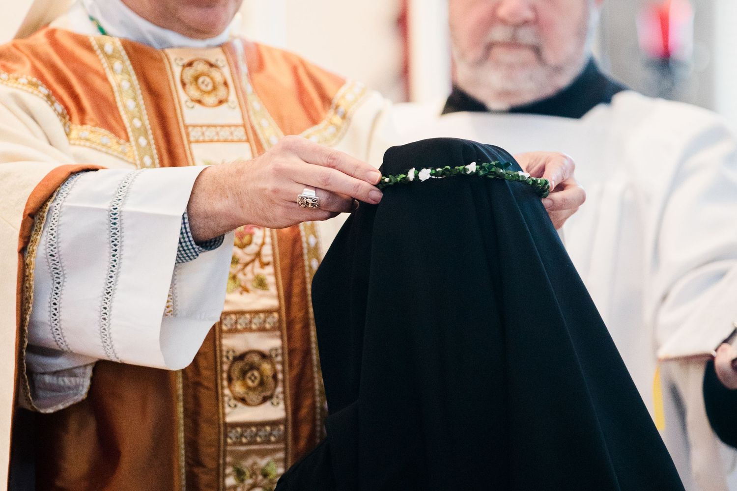 Catholic priest places a floral crown on a nun during a religious vows ceremony with clergy present.