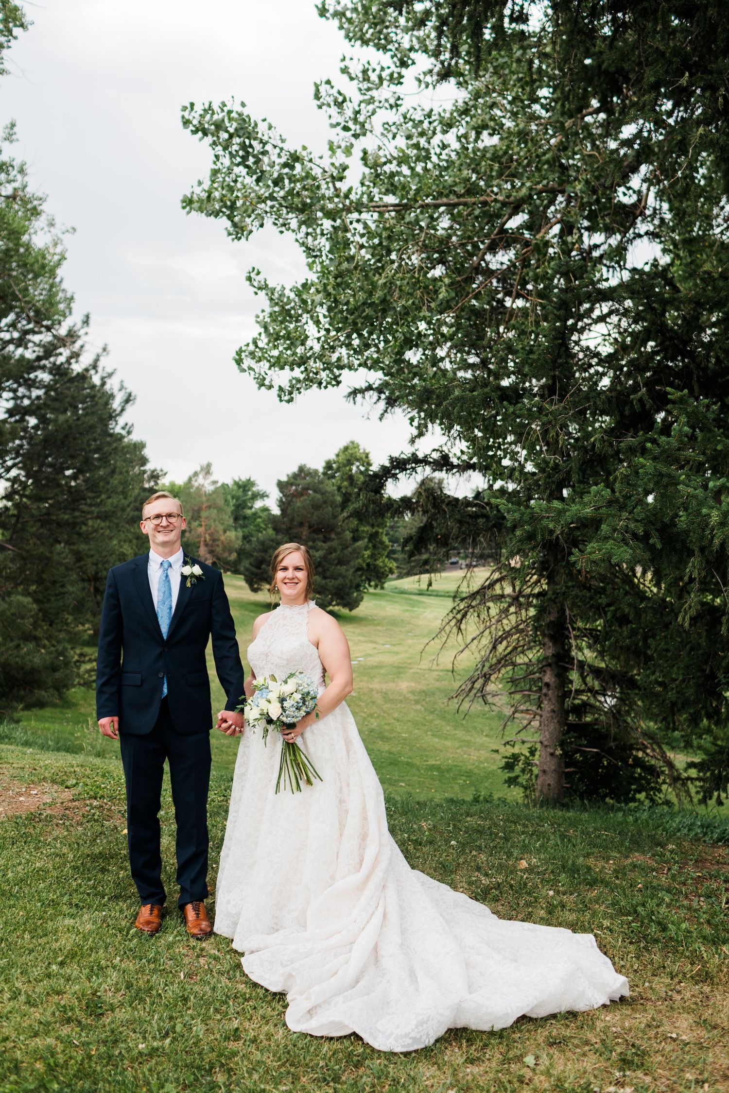 Smiling bride and groom hold hands outdoors on lush green grounds on their wedding day.