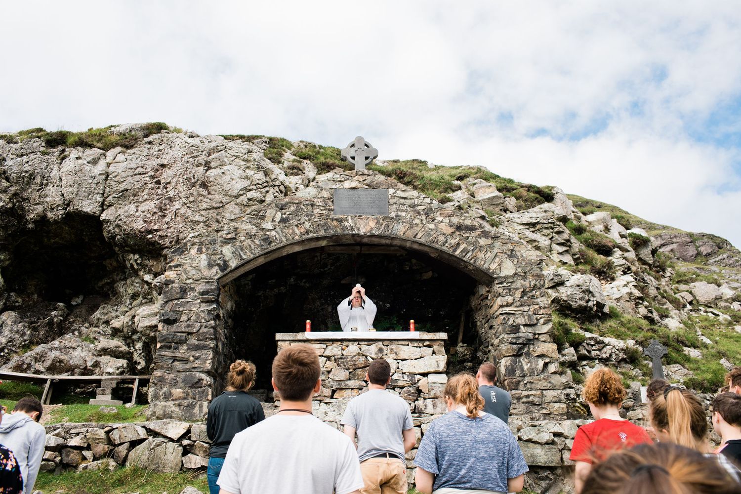 Outdoor mountain Mass celebrated at a stone grotto altar with worshippers gathered before a priest.