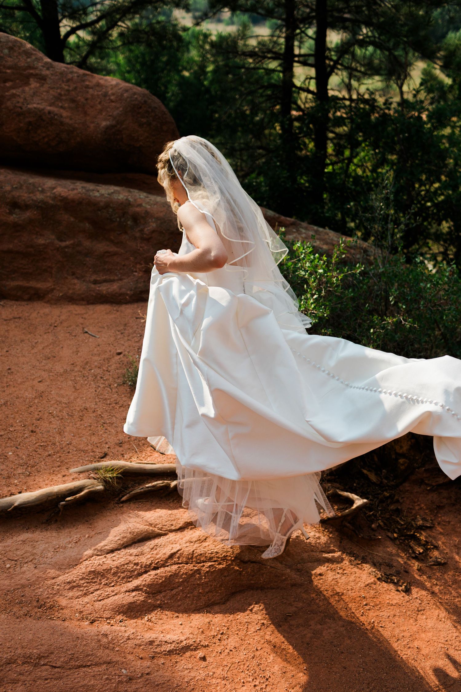 Bride in white wedding gown and veil walking on red rock terrain surrounded by lush greenery.