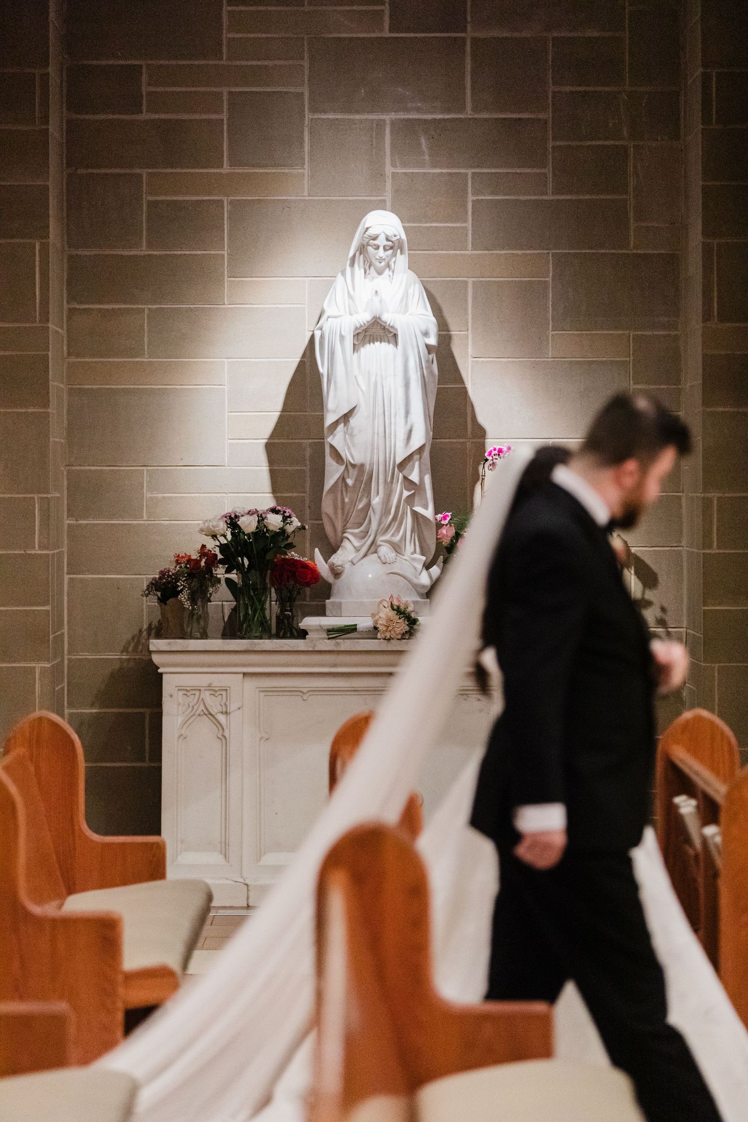 Bride and groom walk past a glowing Virgin Mary statue in a Catholic church during their wedding ceremony.