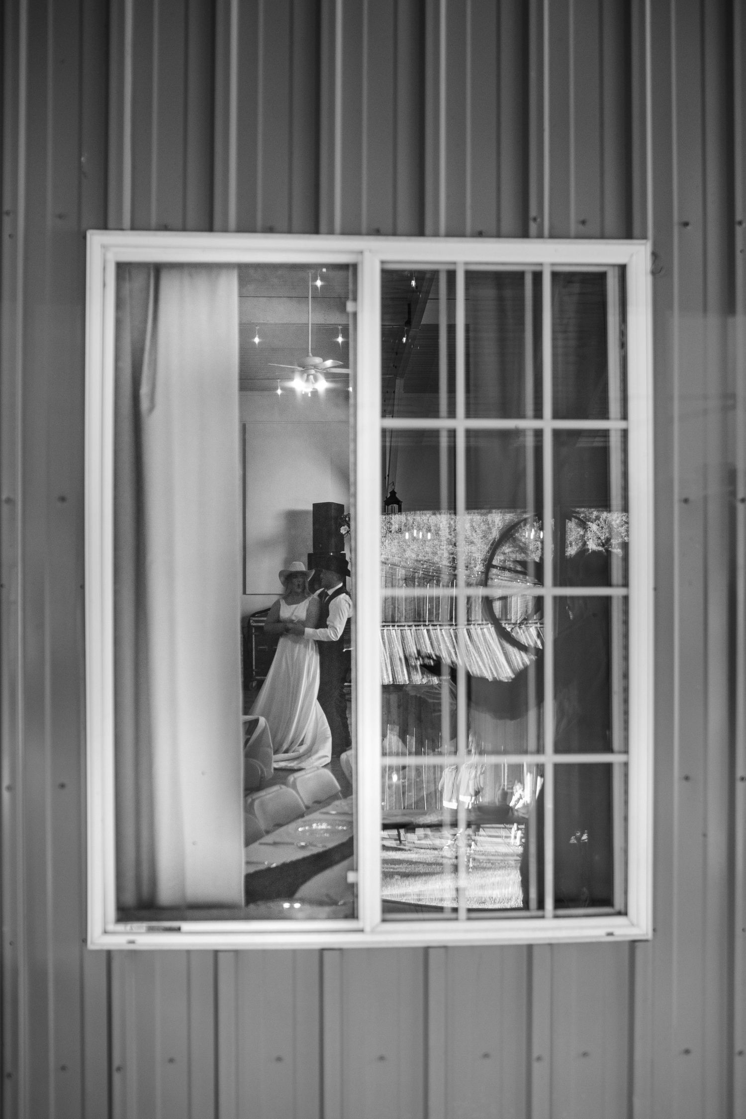 Black and white photo of bride visible through barn window with elegant reception decor inside.