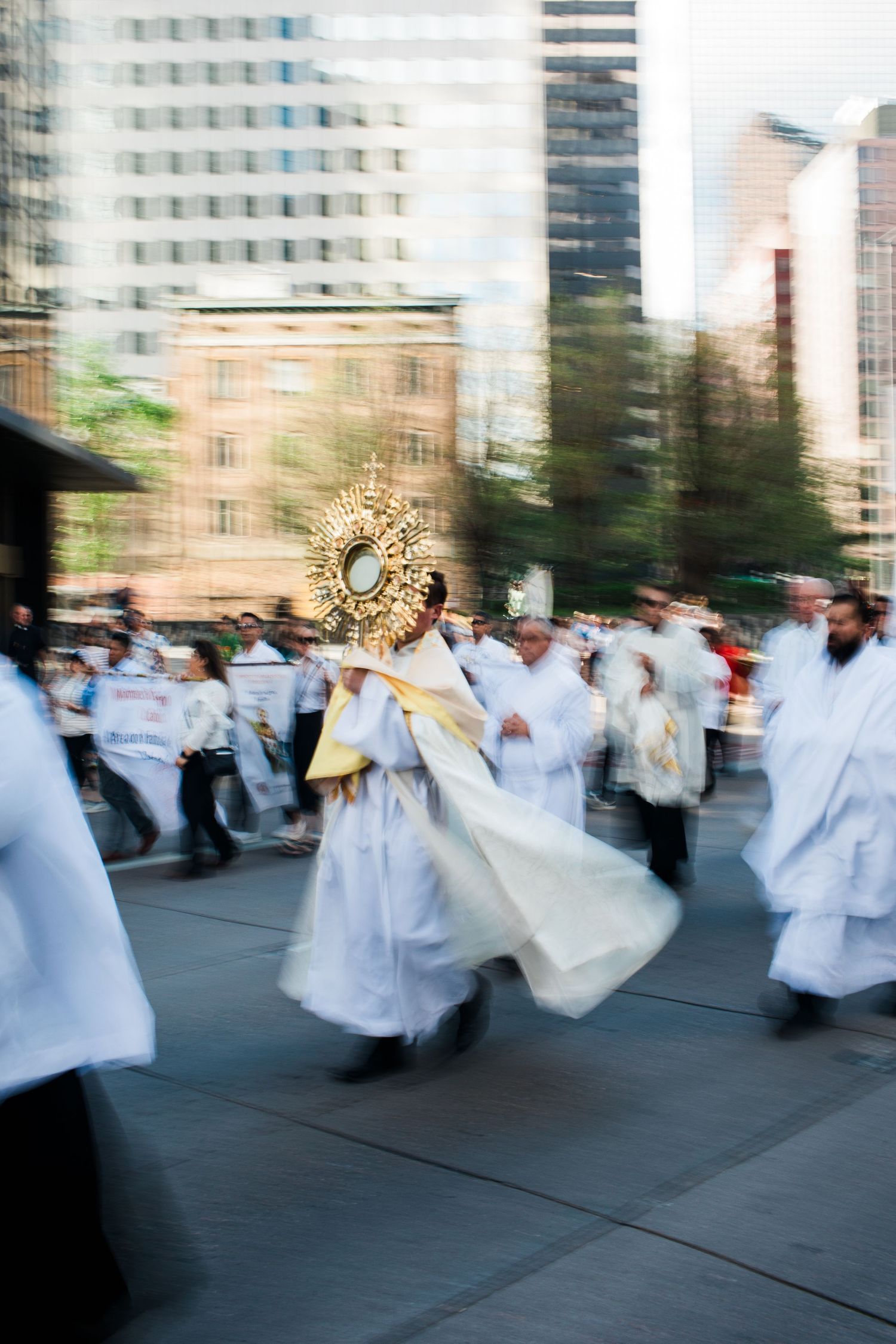 Clergy in white robes carry a golden monstrance in a Corpus Christi procession through a city street.