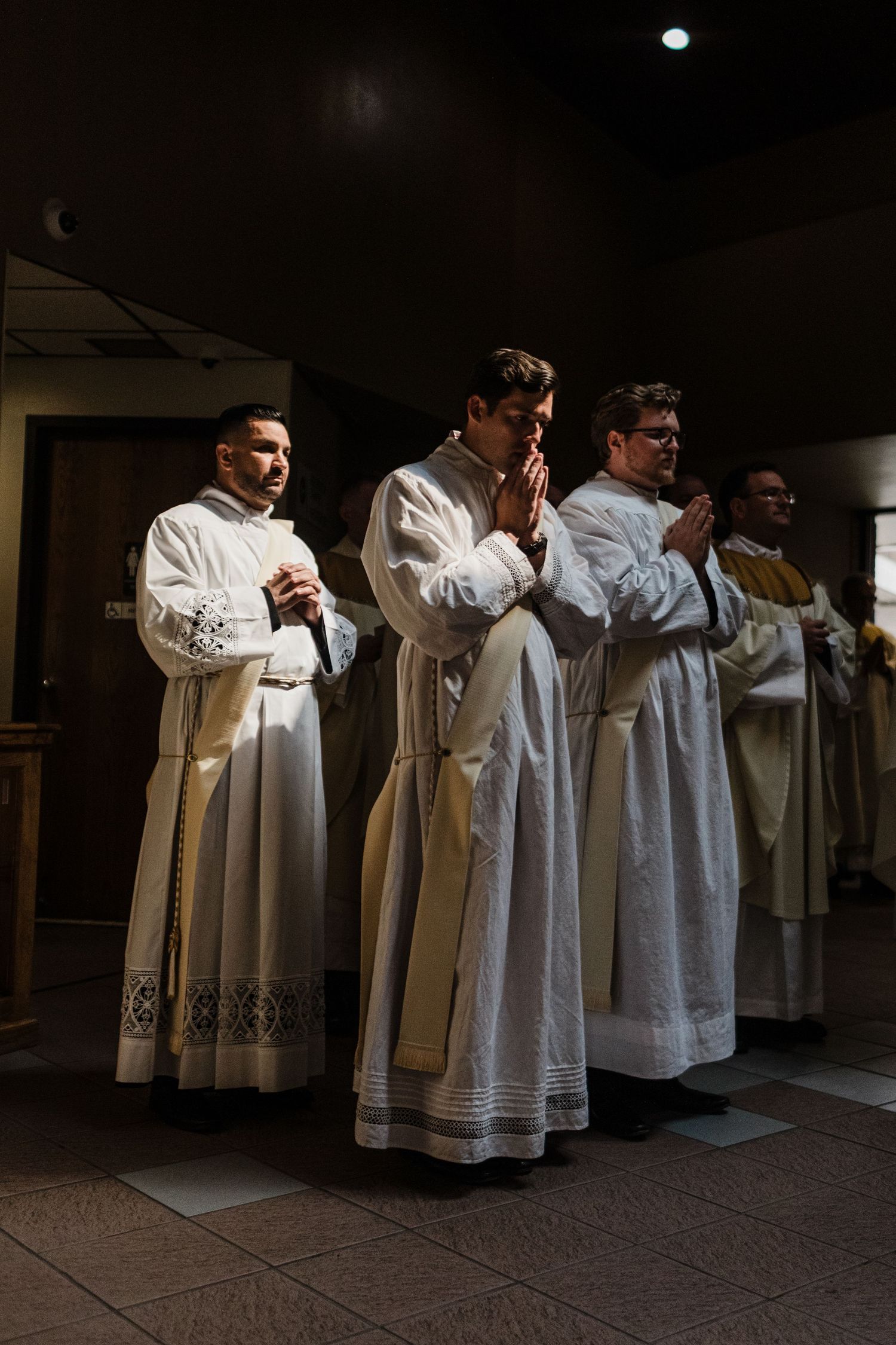 Catholic clergy in white robes pray with hands folded during a religious ceremony inside a church.