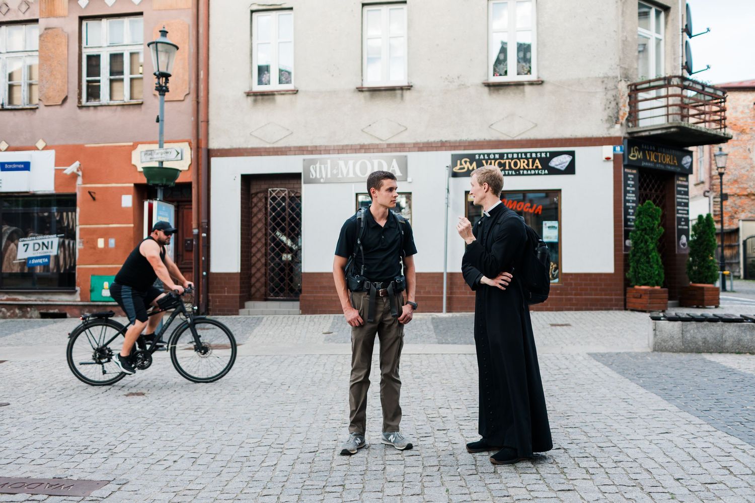 Two men conversing in a European town square, with a cyclist passing by historic buildings in the background.