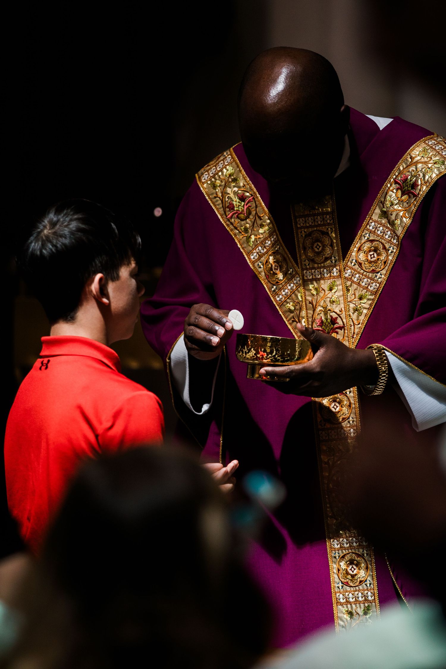Priest wearing ornate purple and gold vestments administers communion to a person in red.