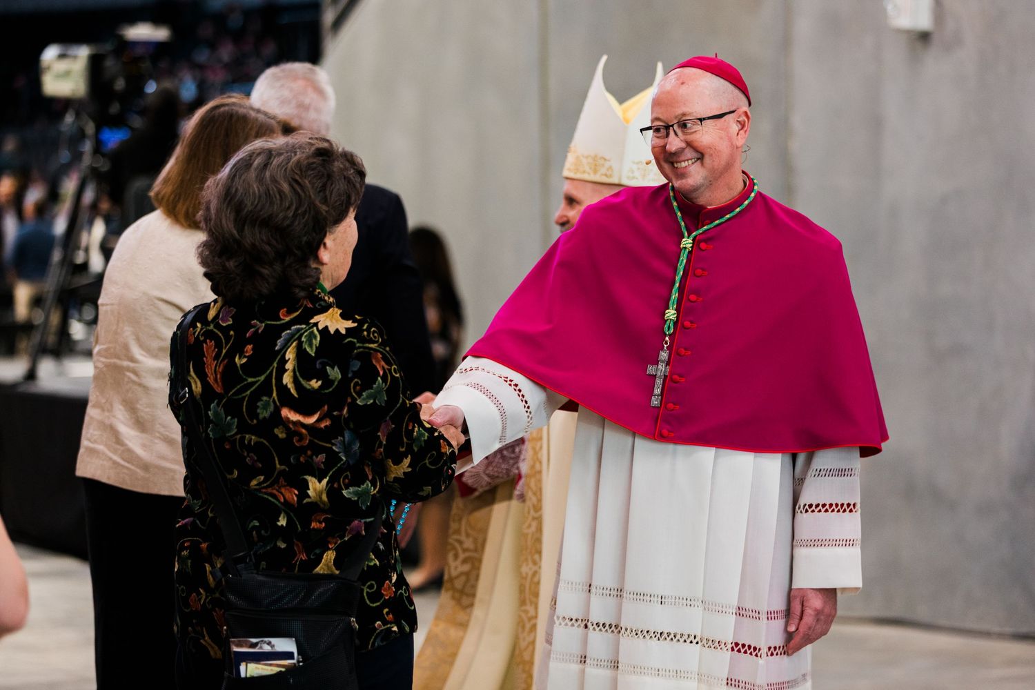 A Catholic bishop in ceremonial magenta robes walks through a church corridor during a formal event.