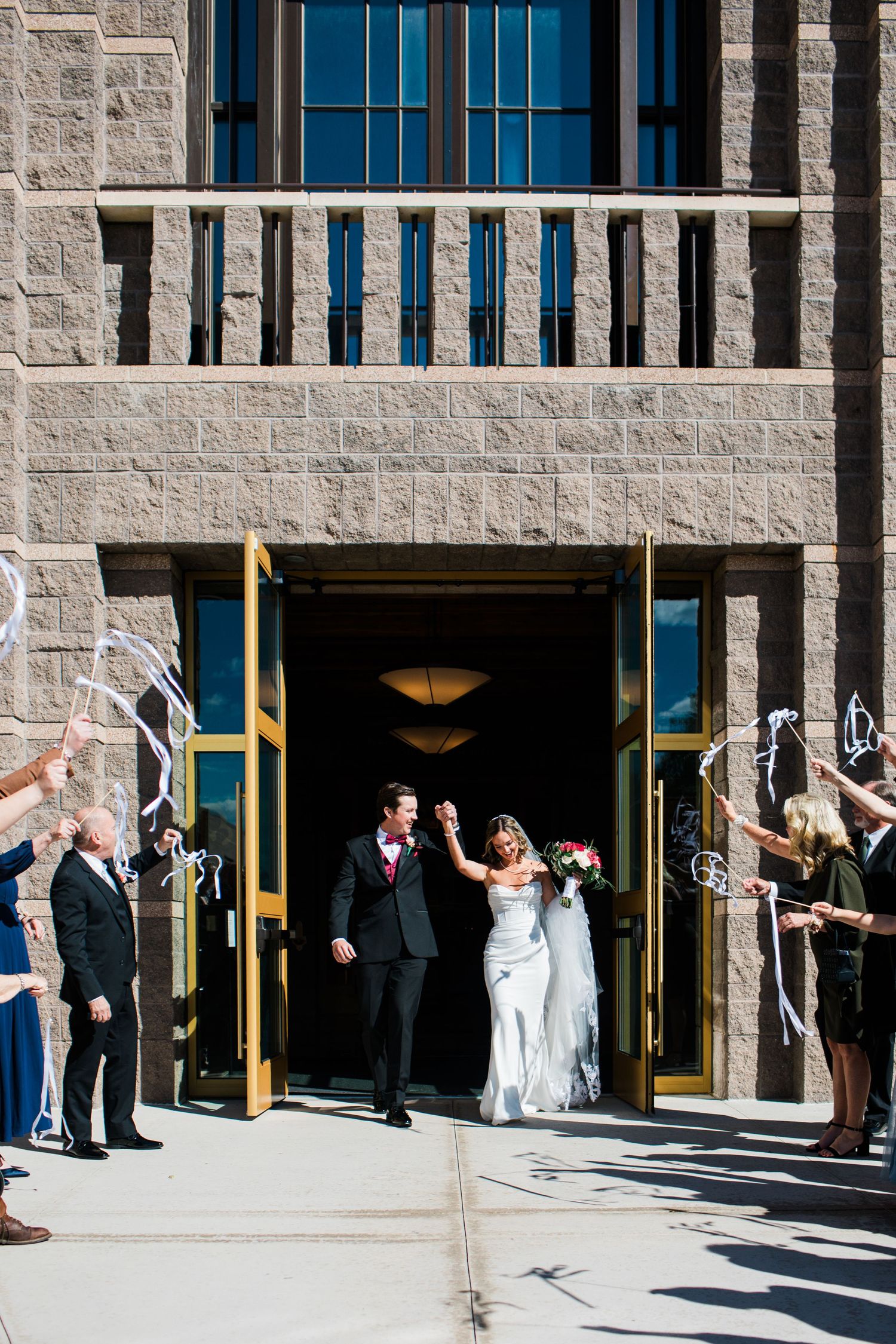 Bride and groom exit church doors holding hands while guests wave ribbon wands in celebration after wedding ceremony.