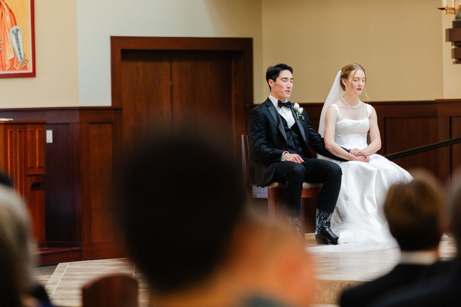 A couple sits together during their wedding ceremony in a church with wooden pews and warm lighting.