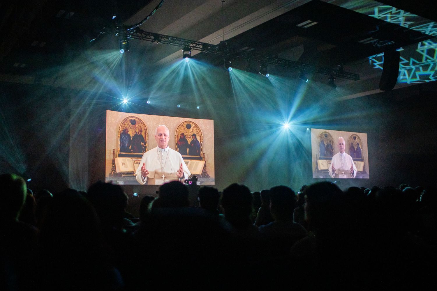 Large concert venue with blue stage lighting and video screens displaying performers during a live performance.