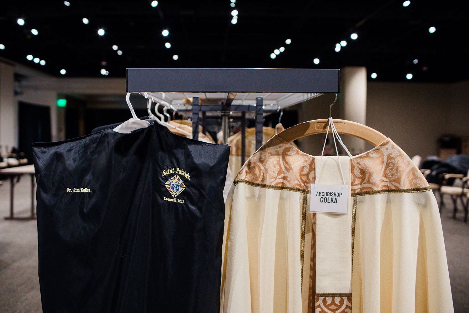 Clergy vestments and garment bags hanging on a rack in a convention hall, including ornate robes and liturgical attire.