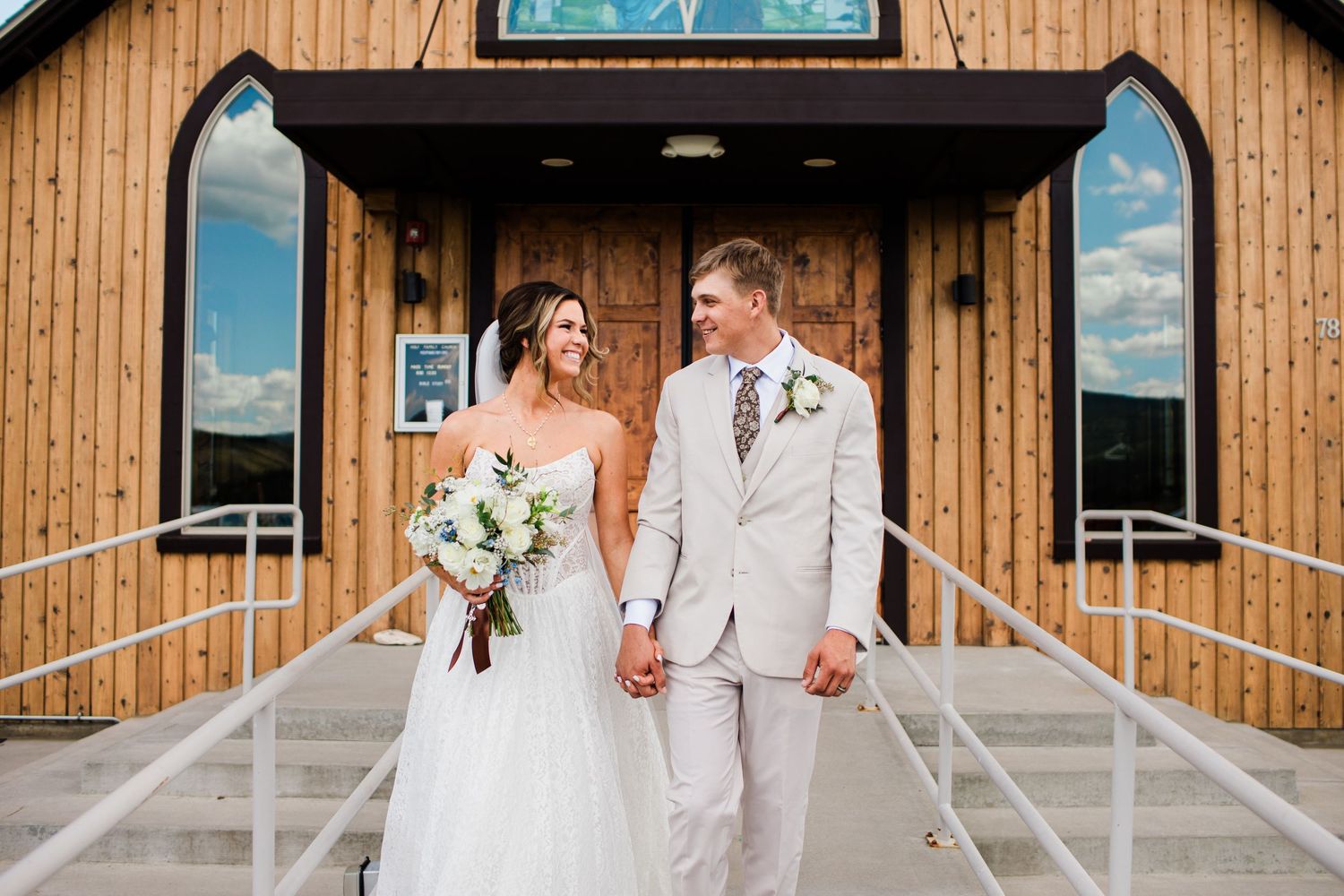 Bride and groom holding hands smiling outside a rustic wooden chapel on their wedding day.