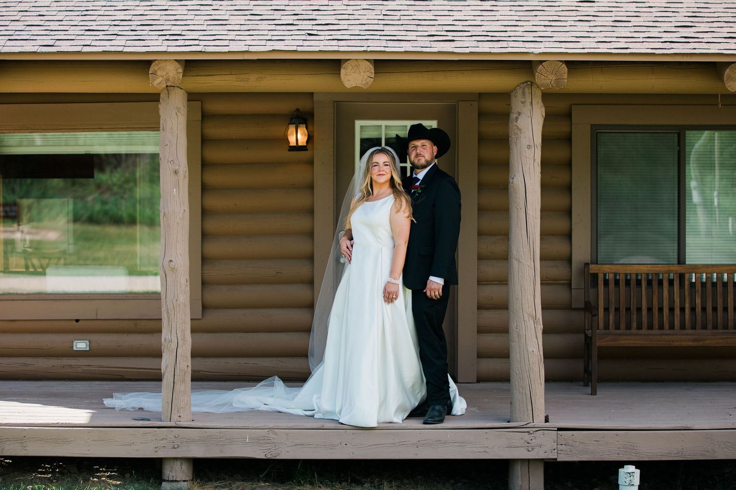 Bride and groom pose on rustic log cabin porch in wedding attire with scenic outdoor backdrop.