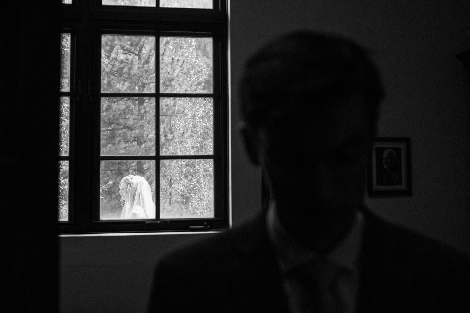 Black and white wedding photo showing groom silhouette indoors with bride in veil visible through window.