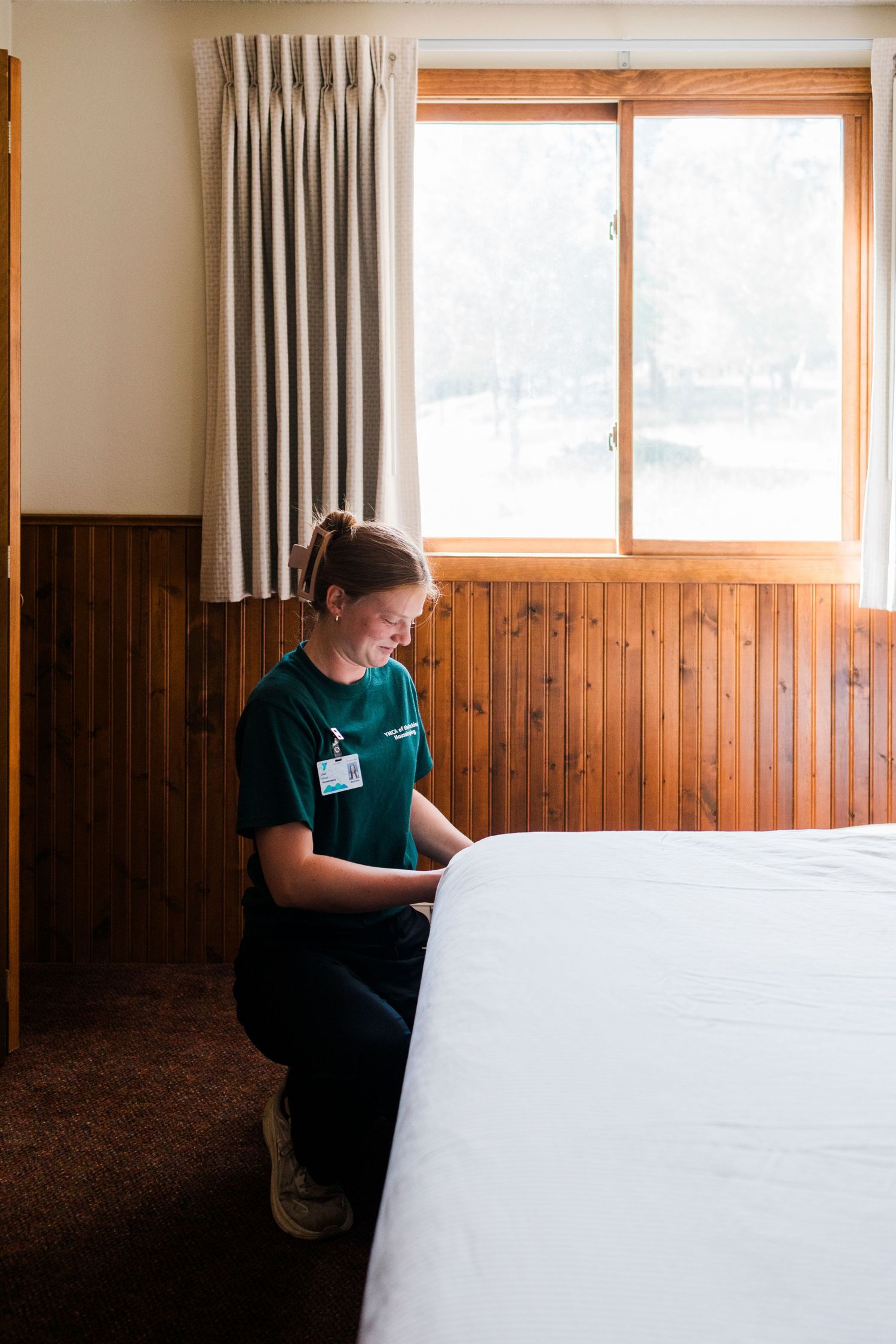Hotel housekeeper in green uniform making a bed in a wood-paneled room with natural window light.