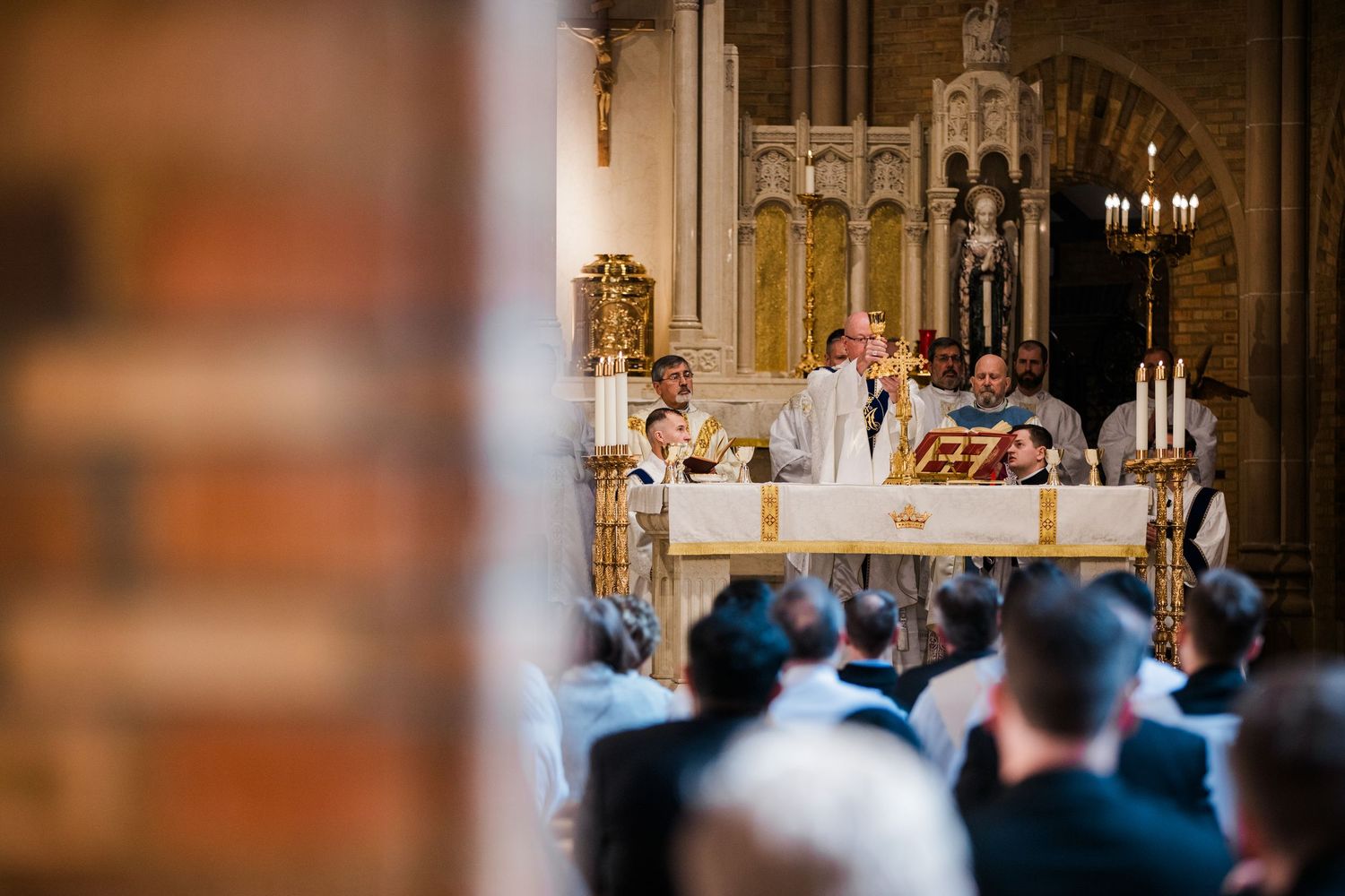 Priest celebrating Catholic Mass at ornate altar with candles, congregation seated in historic church.