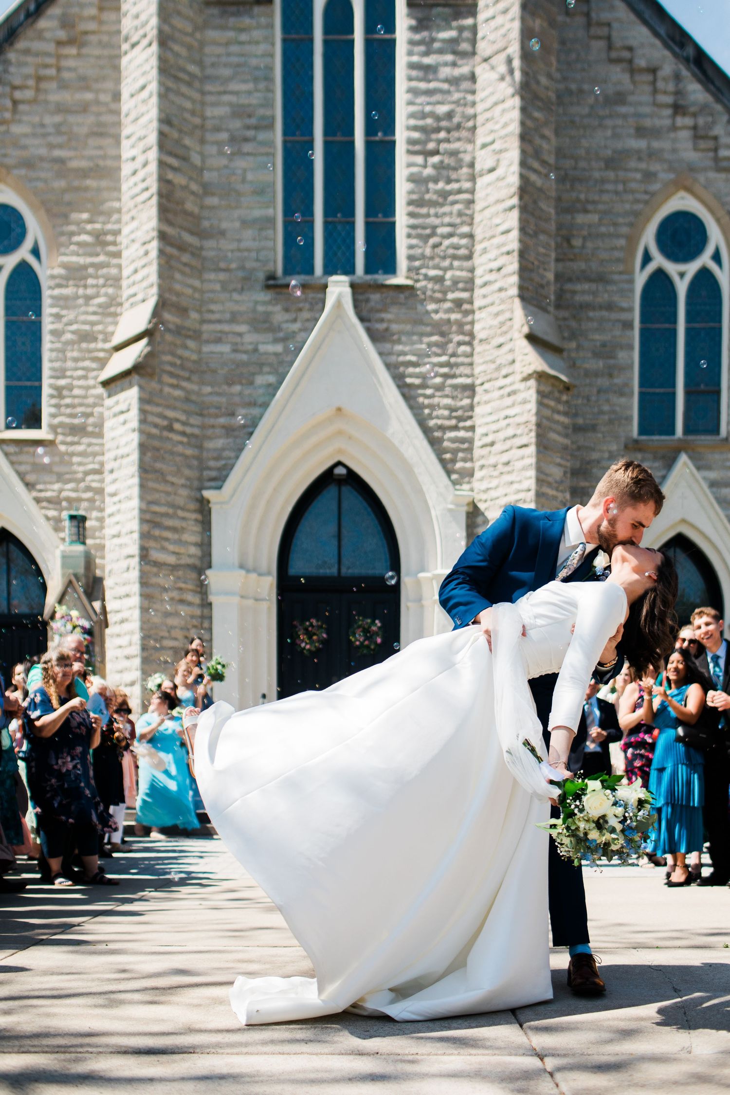 Groom dips bride in kiss outside stone church after wedding ceremony, guests celebrating in background.