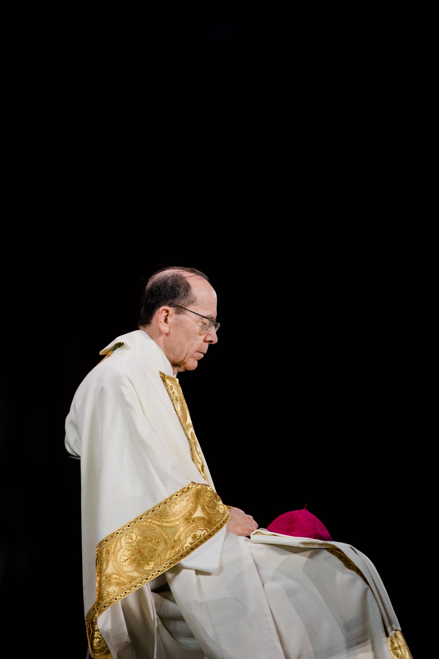 Priest wearing white and gold vestments kneels during a religious ceremony against black background.